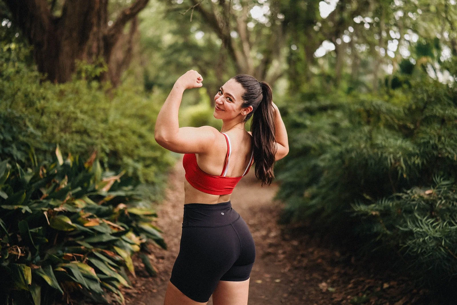 A woman in workout attire confidently flexes her bicep and smiles, standing on a forest trail surrounded by green foliage.