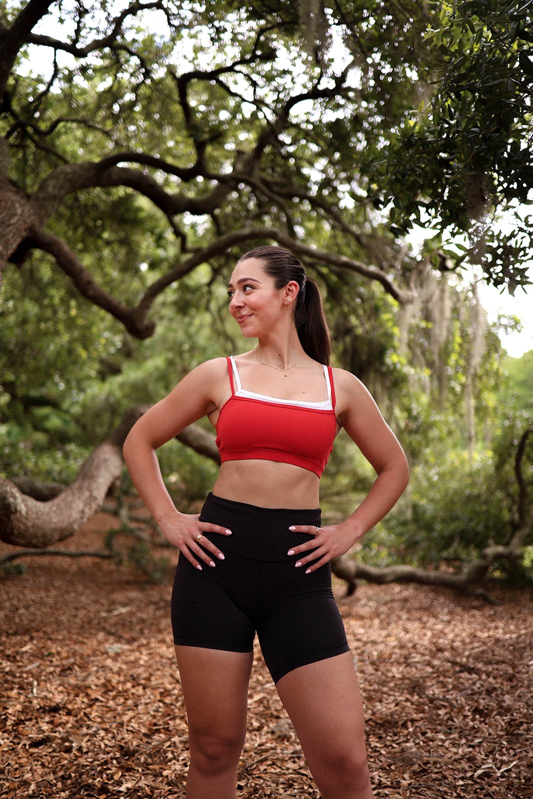 A woman in athletic wear standing outdoors in a wooded area with her hands on her hips, smiling and looking to the side.
