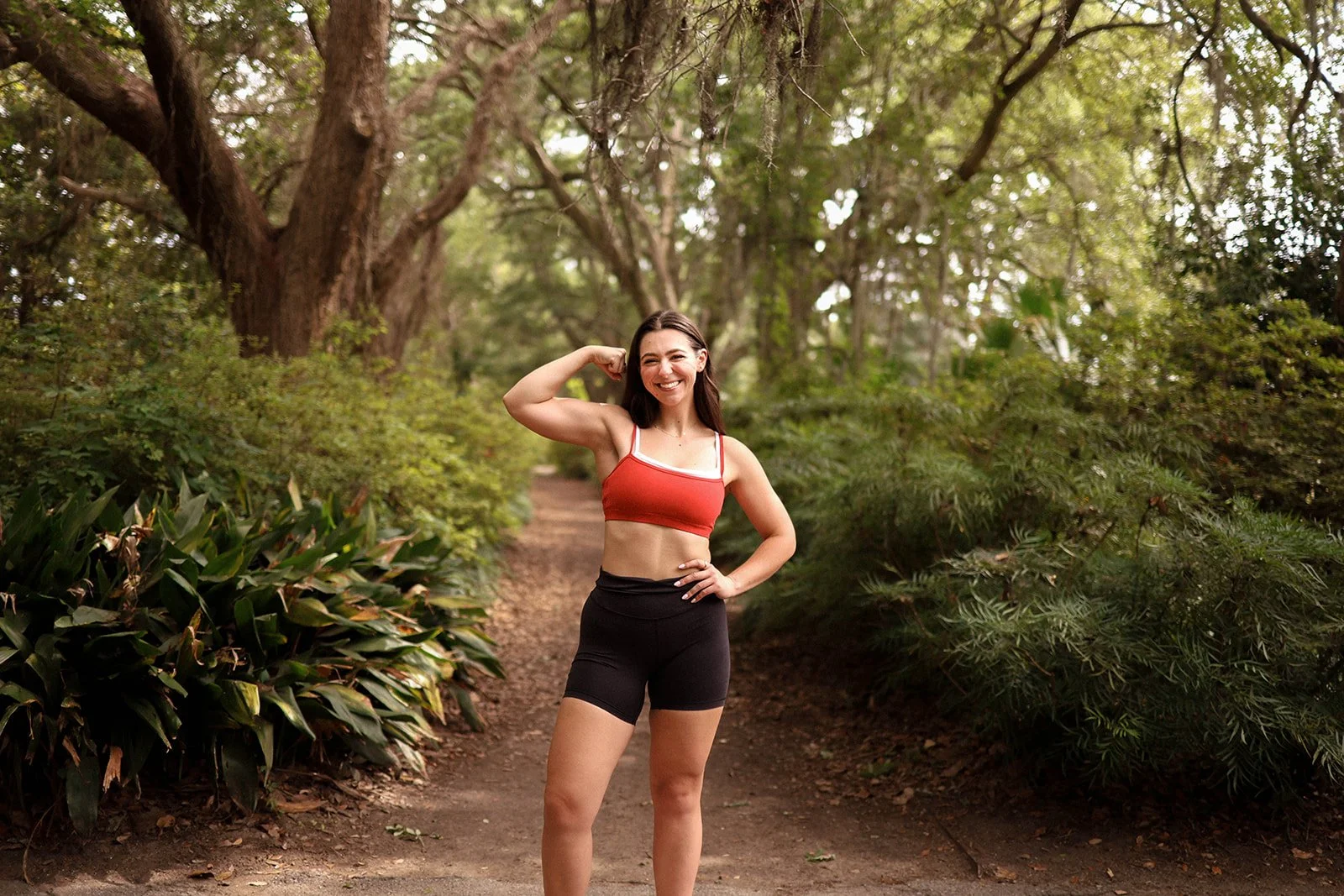 A woman in athletic clothing posing with a flexed arm muscle in a wooded park.