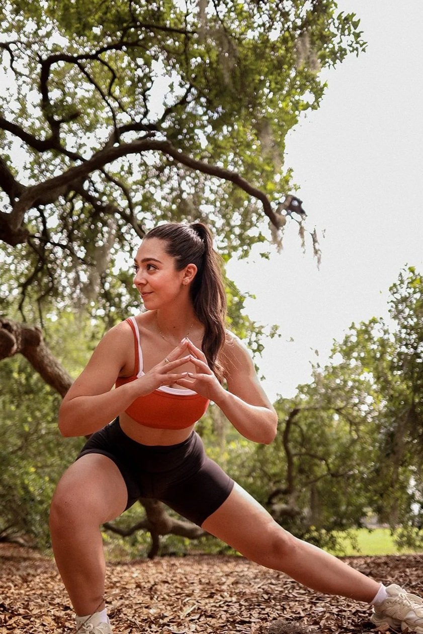 A woman practicing yoga outdoors, squatting with her hands in a prayer position, surrounded by trees and fallen leaves.