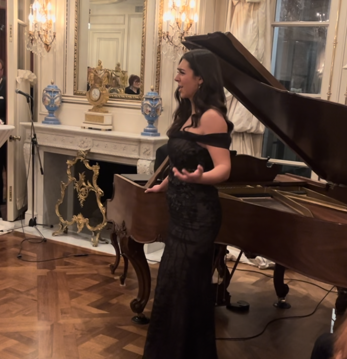 A woman in an elegant black dress standing beside a grand piano in a luxurious, ornately decorated room, holding a small object in her right hand and smiling.