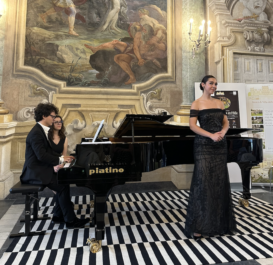 A woman in a black lace off-the-shoulder gown sings while a man and woman play a grand piano together in an ornate, historic room with a large mural painting and decorative wall features.