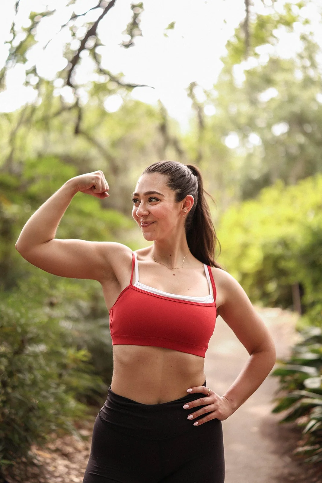 A woman standing outdoors on a wooded path, wearing a red sports bra and black leggings, flexing her right arm and smiling.