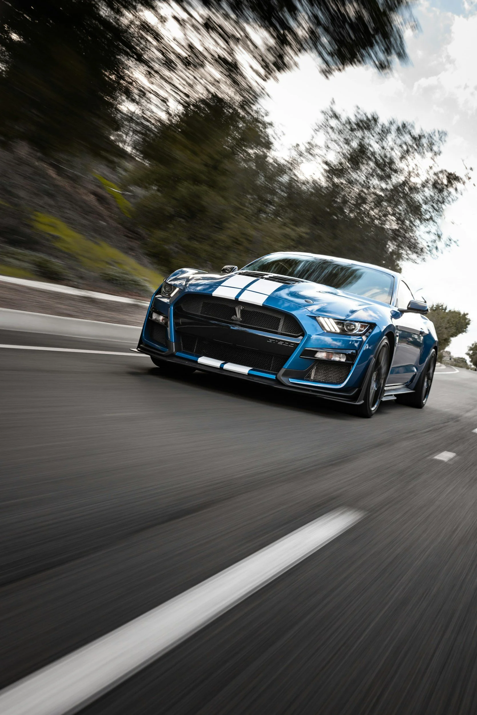 A blue sports car with white racing stripes driving fast on a curved road with trees and sky in the background.