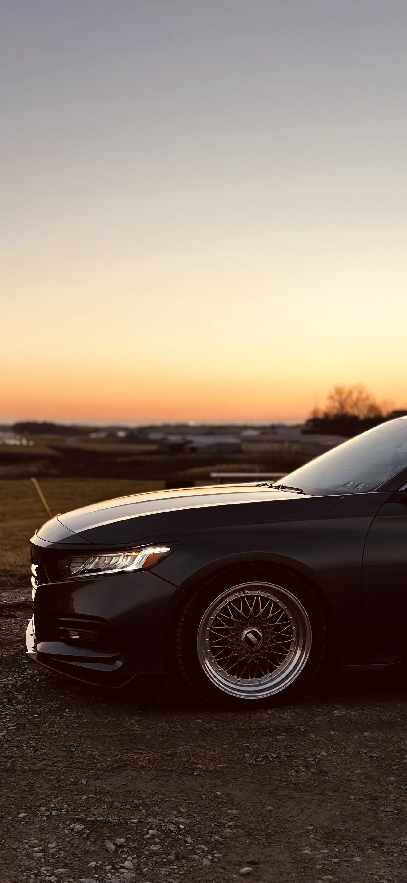 Close-up of a black car with wire-spoke wheels parked on a gravel surface during sunset, with a landscape and a colorful sky in the background.