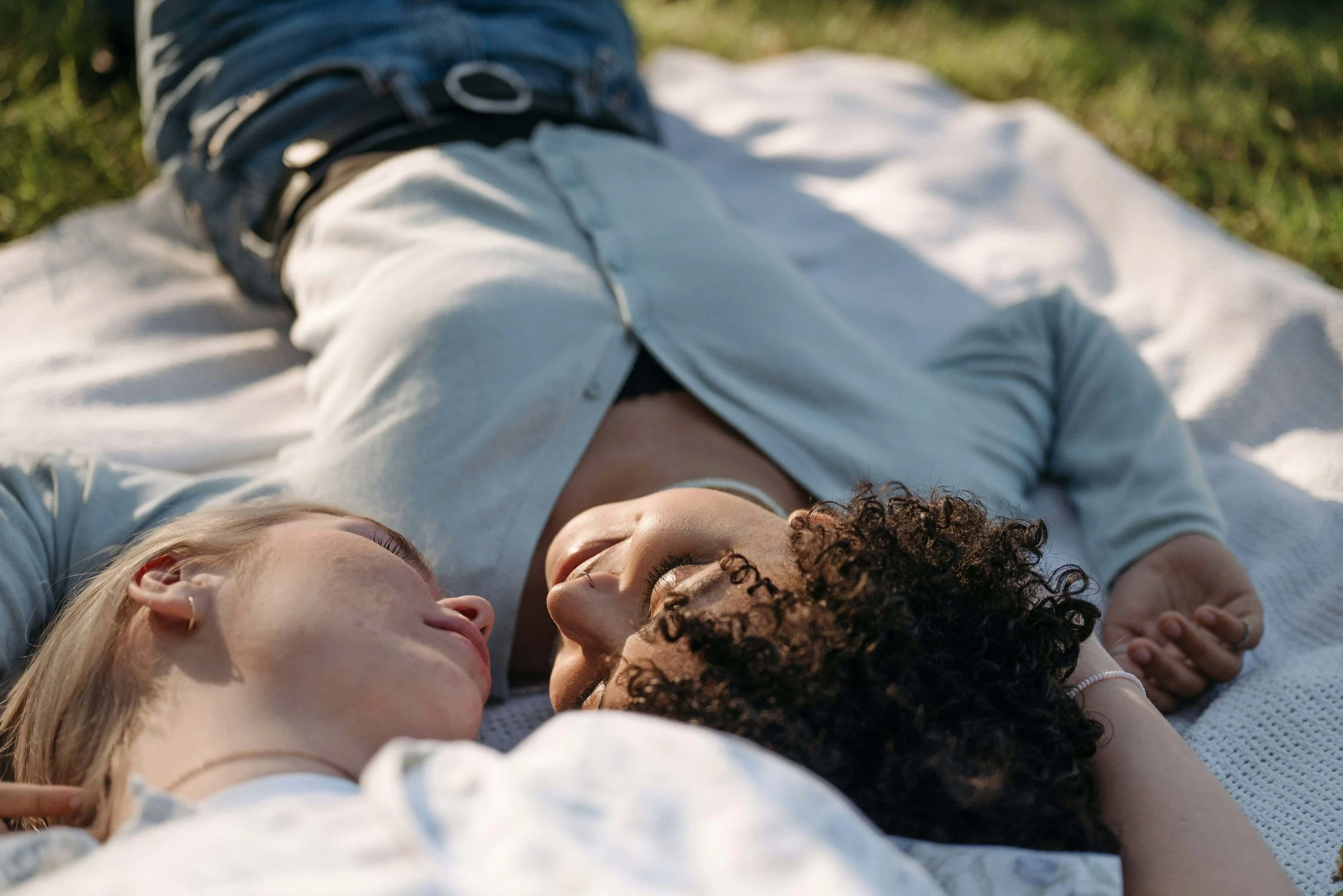 Two women lying on a white blanket outdoors, facing each other and smiling, with closed eyes, surrounded by green grass.