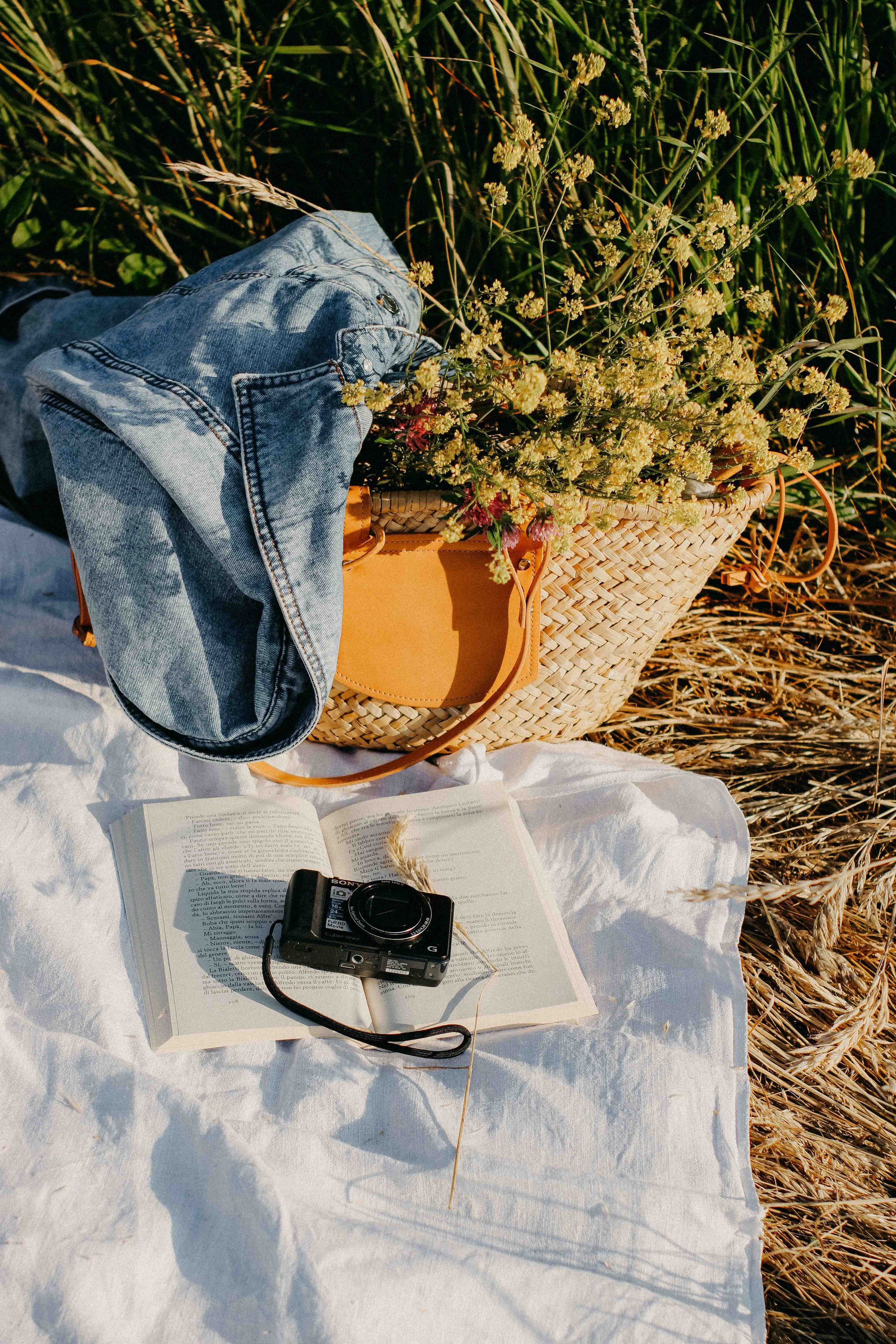 A wicker basket filled with yellow flowers is placed on a white cloth on the ground amidst tall grass. An open book and a black camera with a strap are also on the cloth. The scene appears to be outdoors, possibly during late afternoon or early evening.