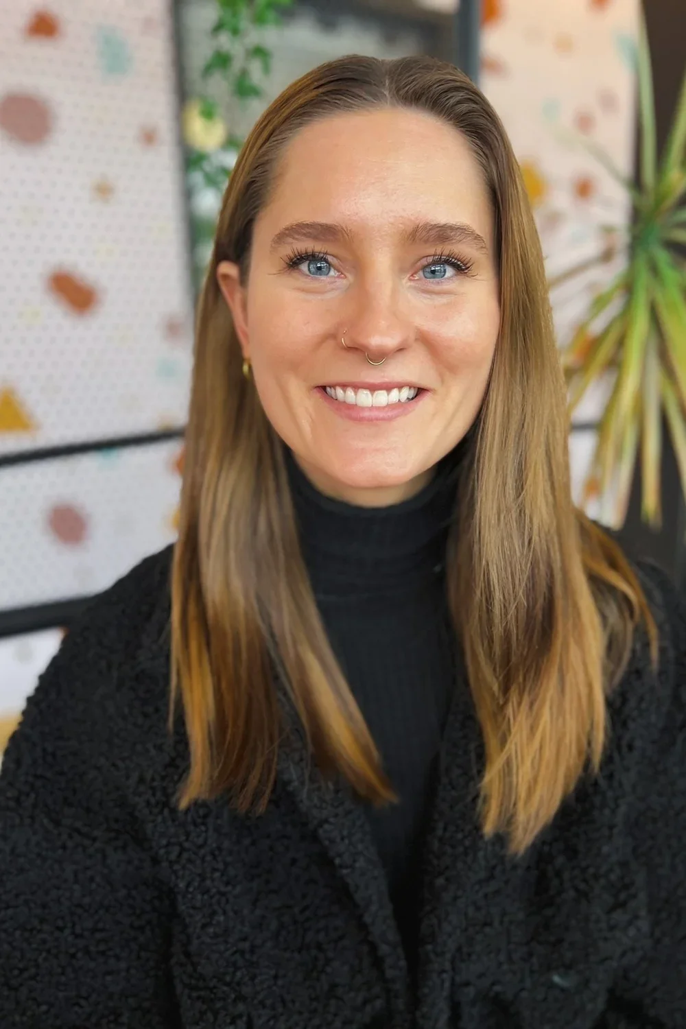 A young person with long light brown hair, blue eyes, and a nose ring smiling, wearing a black turtleneck and black textured sweater, with colorful plants and a pegboard in the background