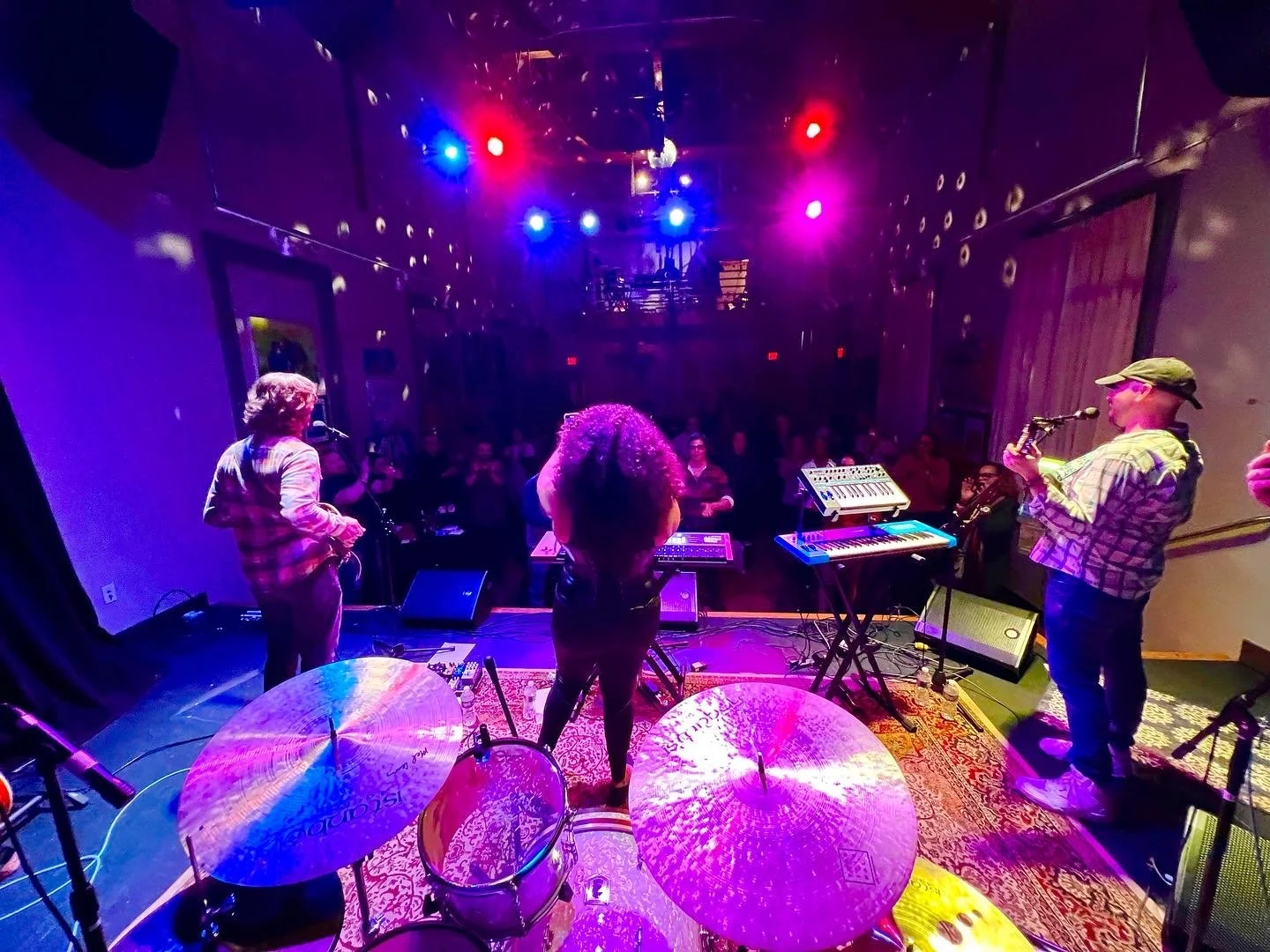 Live music performance at an indoor venue with colorful stage lights. Musicians are playing instruments, and audience members are watching.