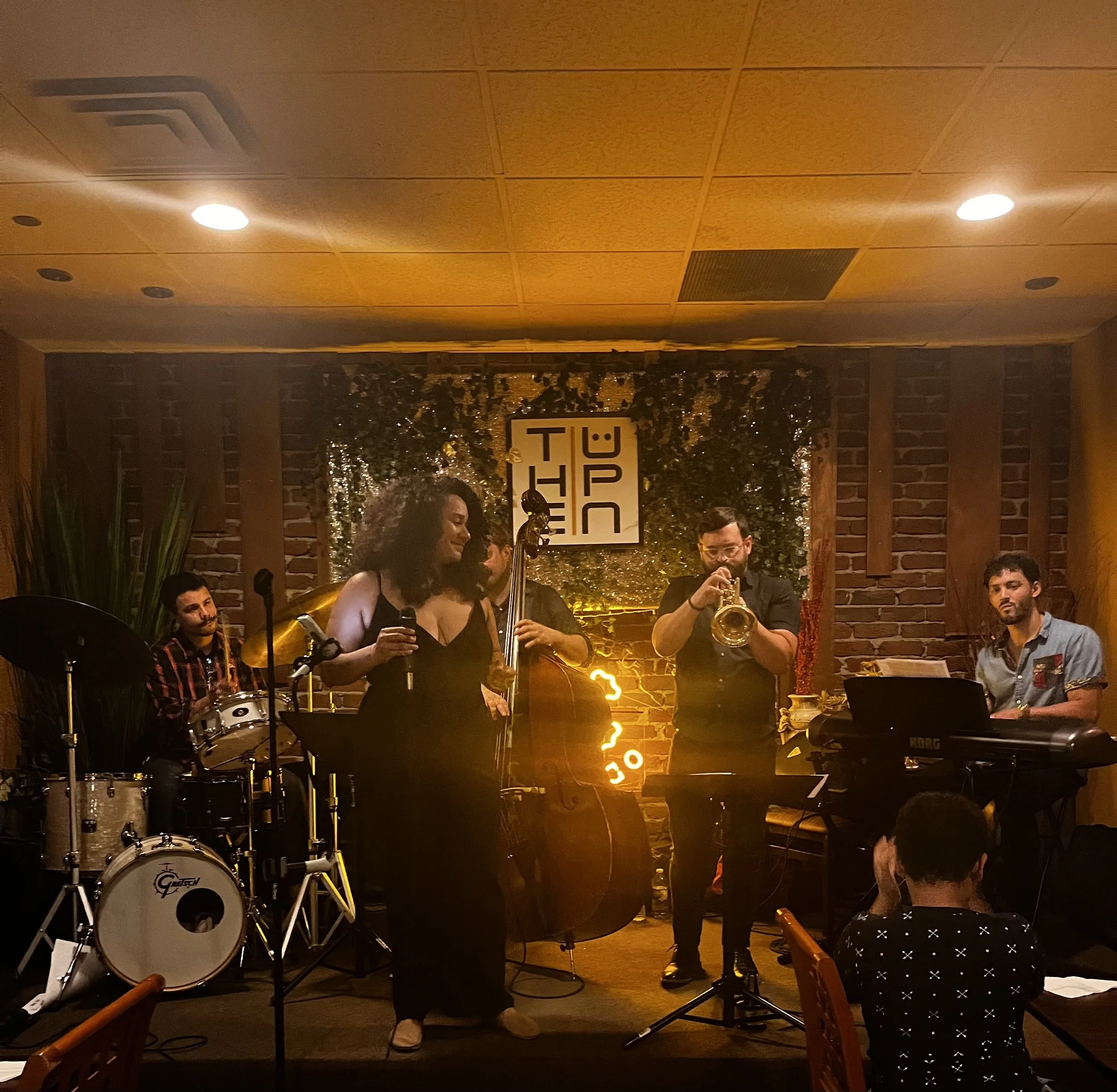 Jazz band performing indoors at night with warm lighting, brick wall, and a sign that reads 'THE T'.