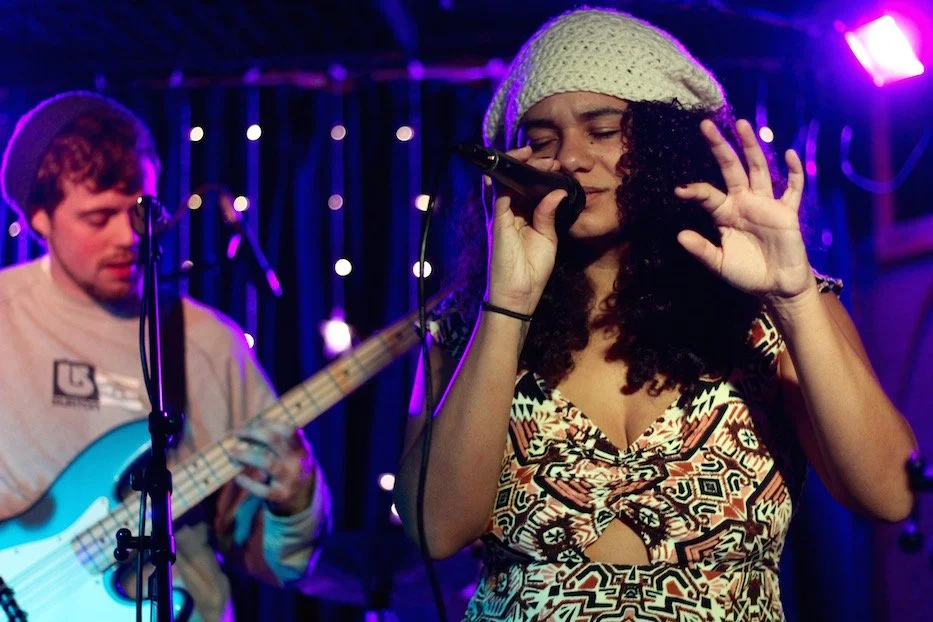A woman with curly hair, wearing a beige knit hat and patterned dress, singing into a microphone during a live music performance with a male guitarist in the background, under purple stage lighting.