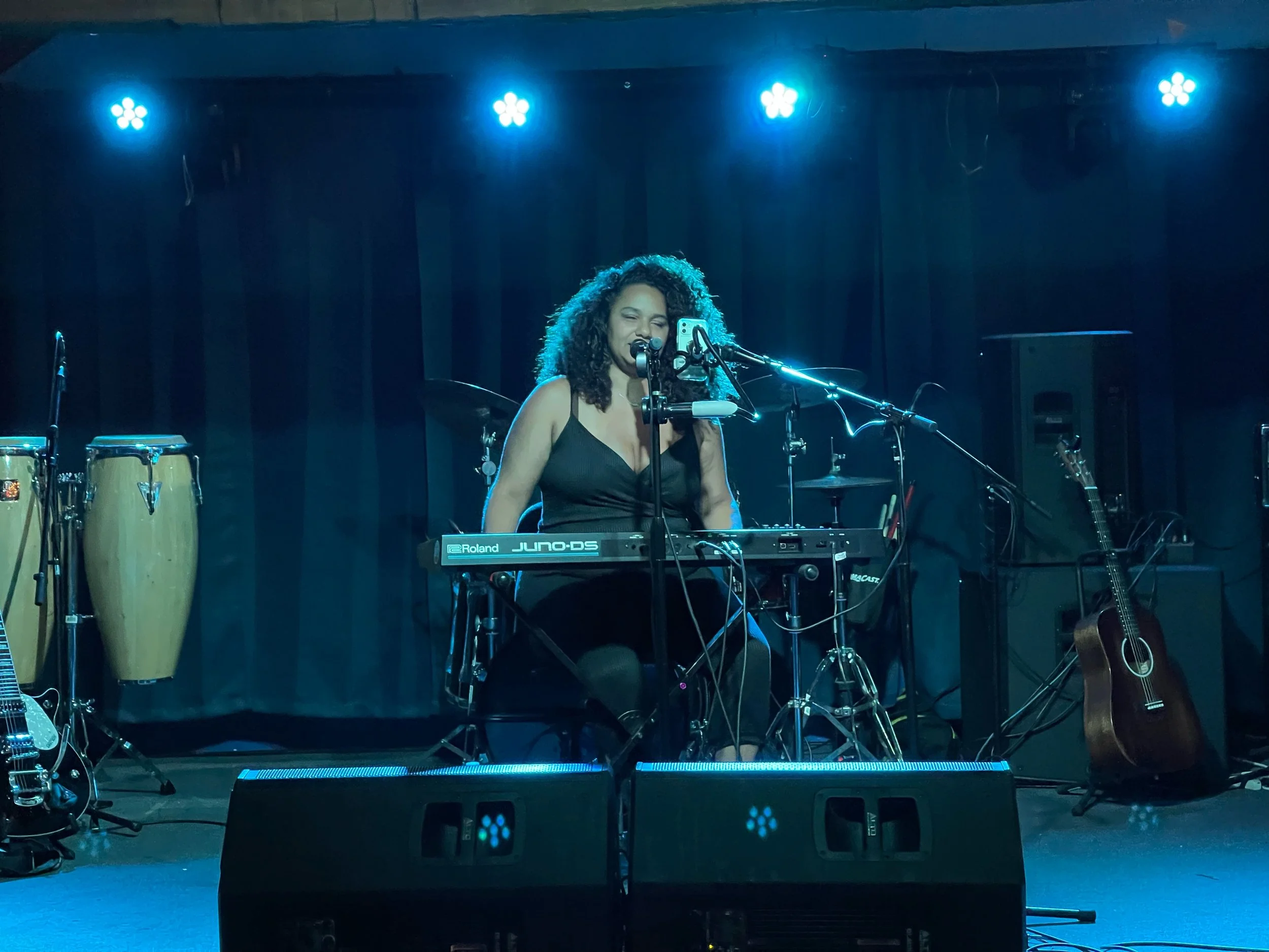 A woman with curly hair singing into a microphone behind a keyboard on stage, with blue stage lights overhead. Musical instruments including congas, drums, and a guitar are visible in the background.