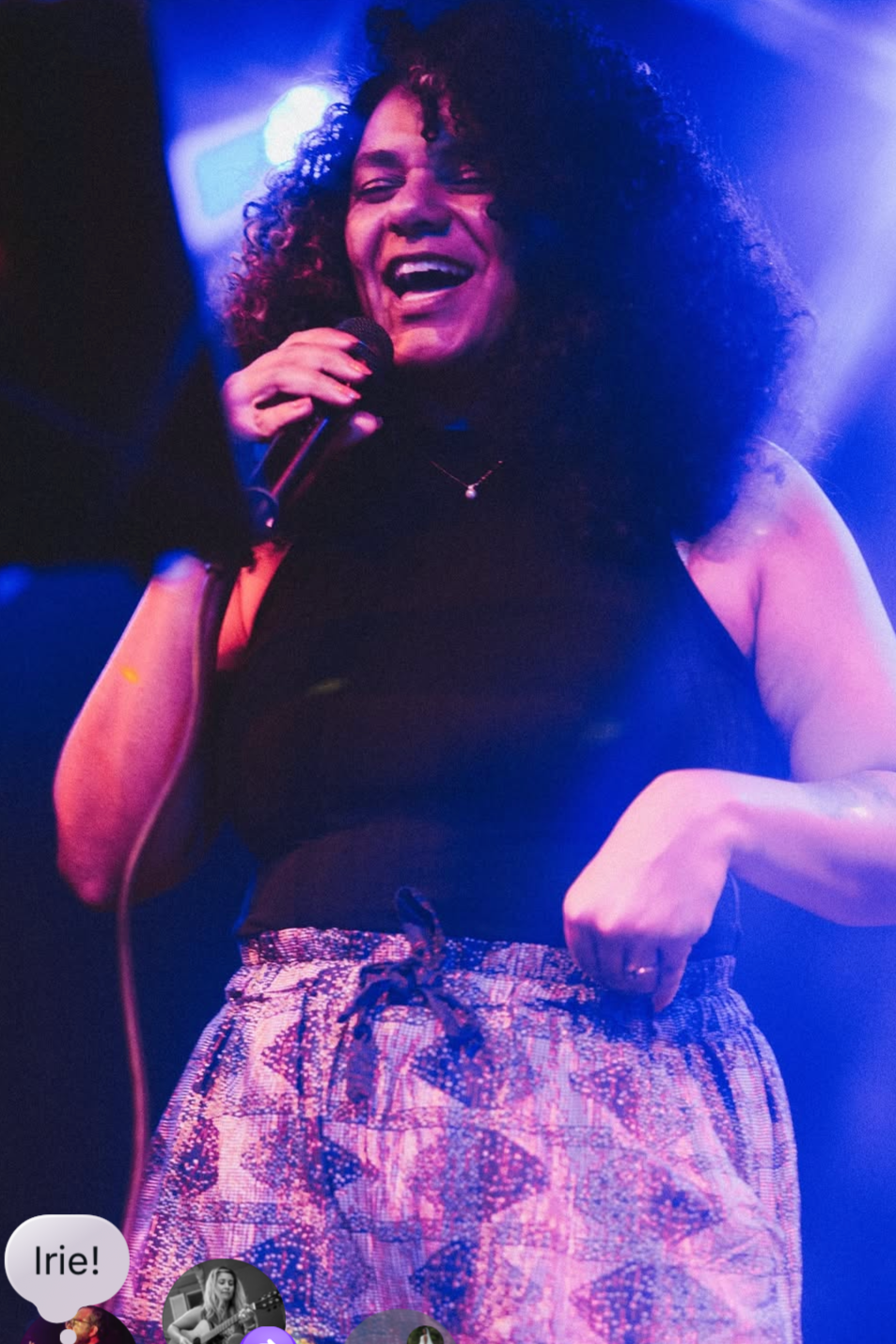 A woman with curly hair singing into a microphone, smiling, under colorful stage lights.