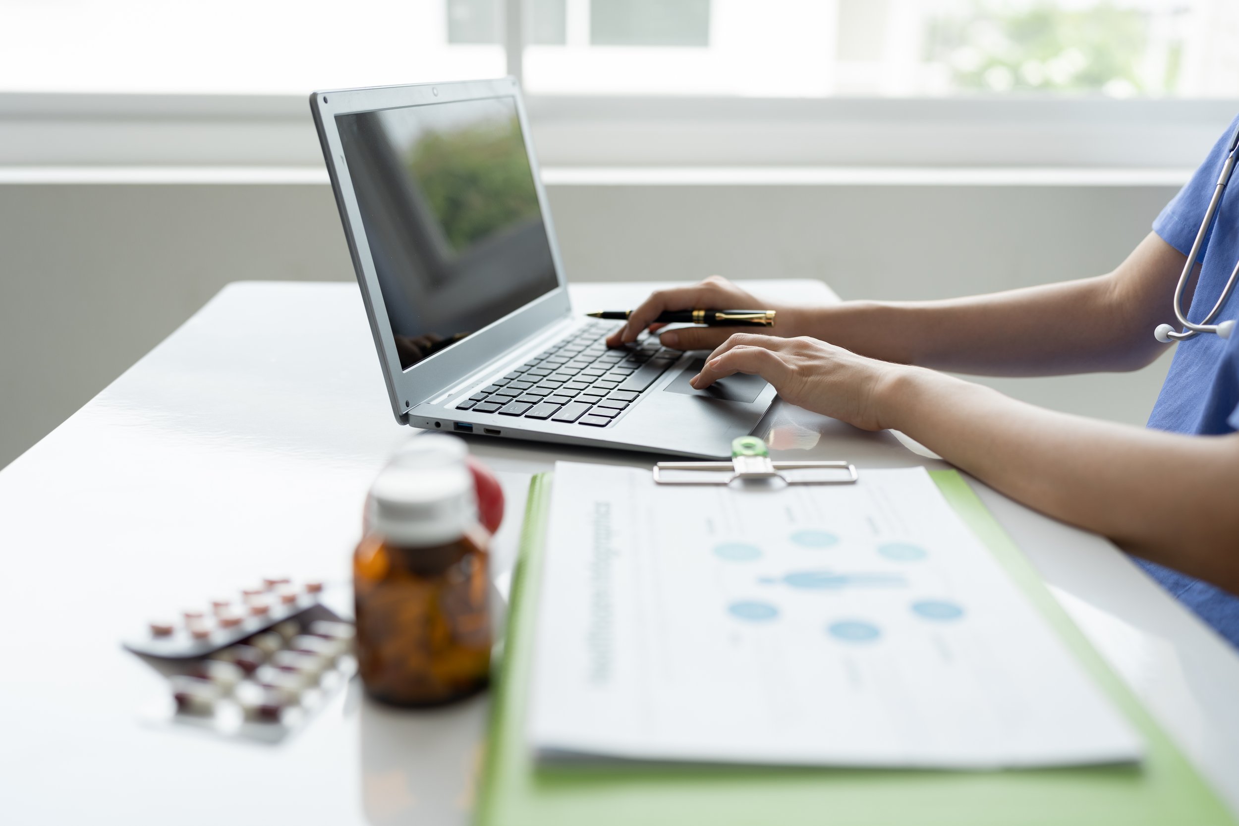 Person in medical scrubs using a laptop computer on a desk with medication bottles and a clipboard nearby in a bright room.