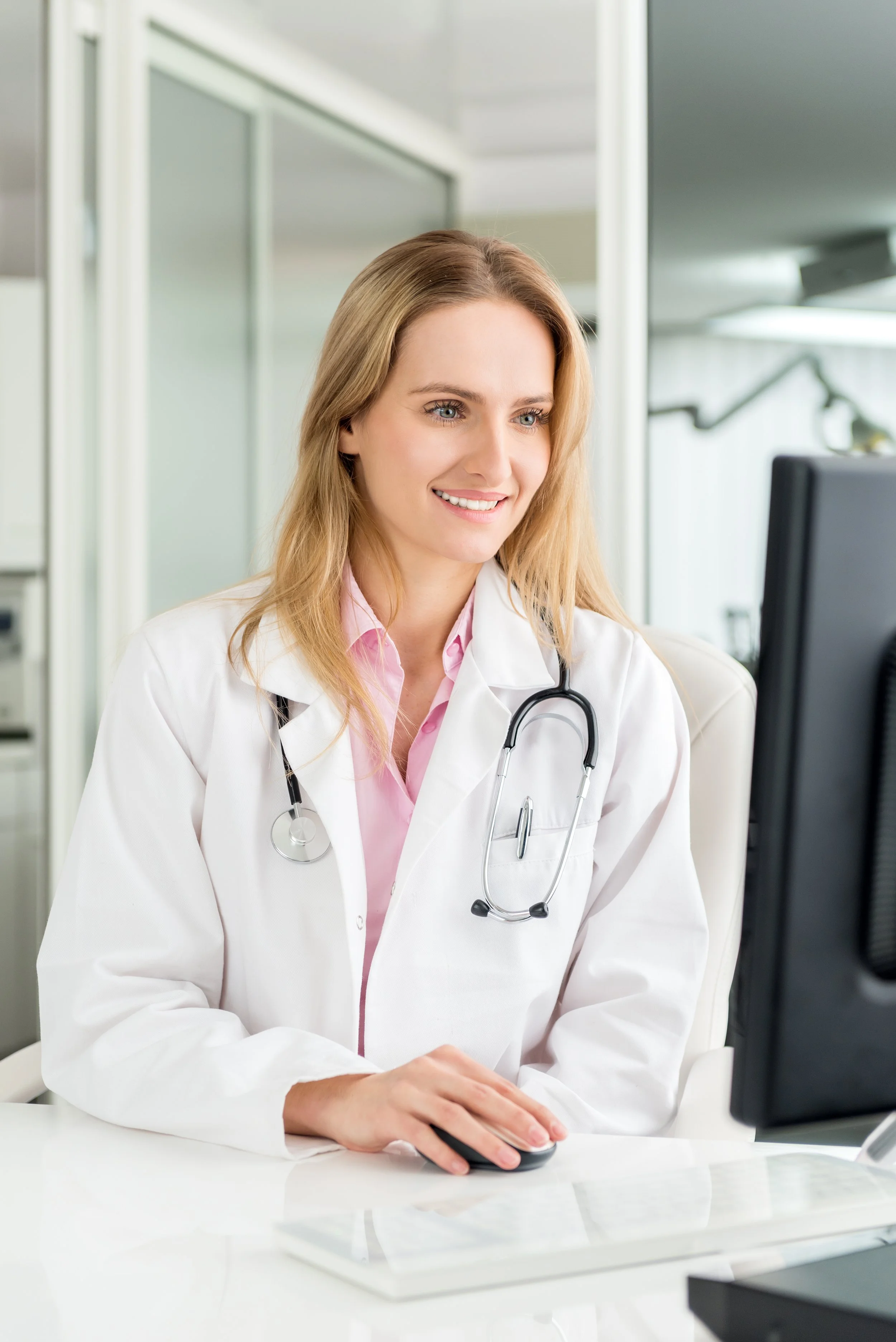 A female doctor with blonde hair and blue eyes, wearing a white medical coat and stethoscope, sitting at a desk and using a computer in a medical office.