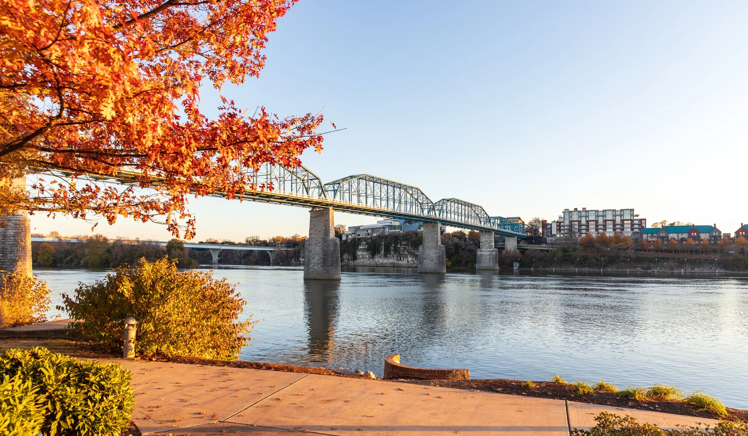 An autumn scene of a river with a bridge crossing over it, framed by colorful fall foliage on the left and in the foreground, with a clear sky in the background.