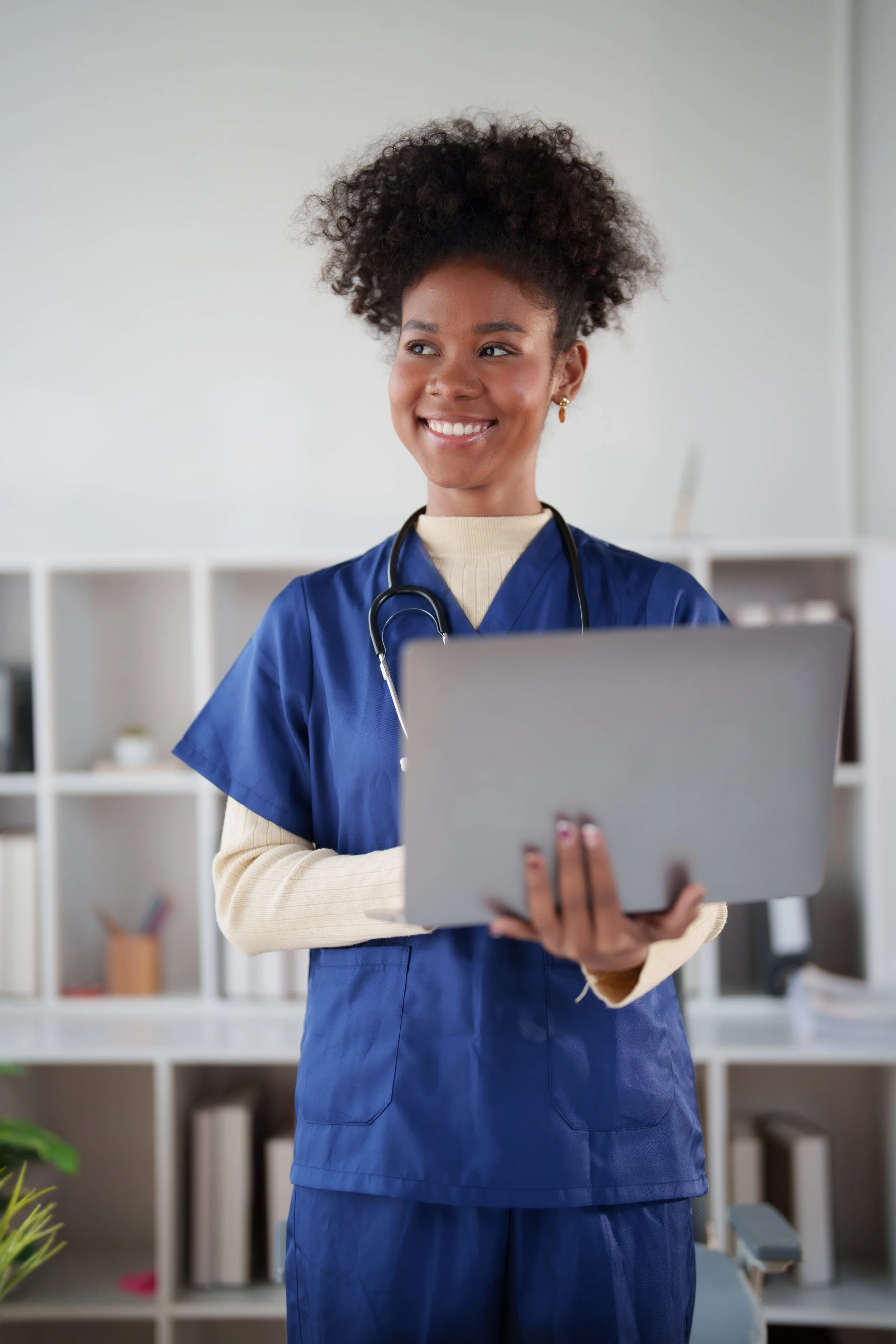 A smiling young woman in blue medical scrubs holding a tablet, standing in front of a white bookshelf with decorative items.