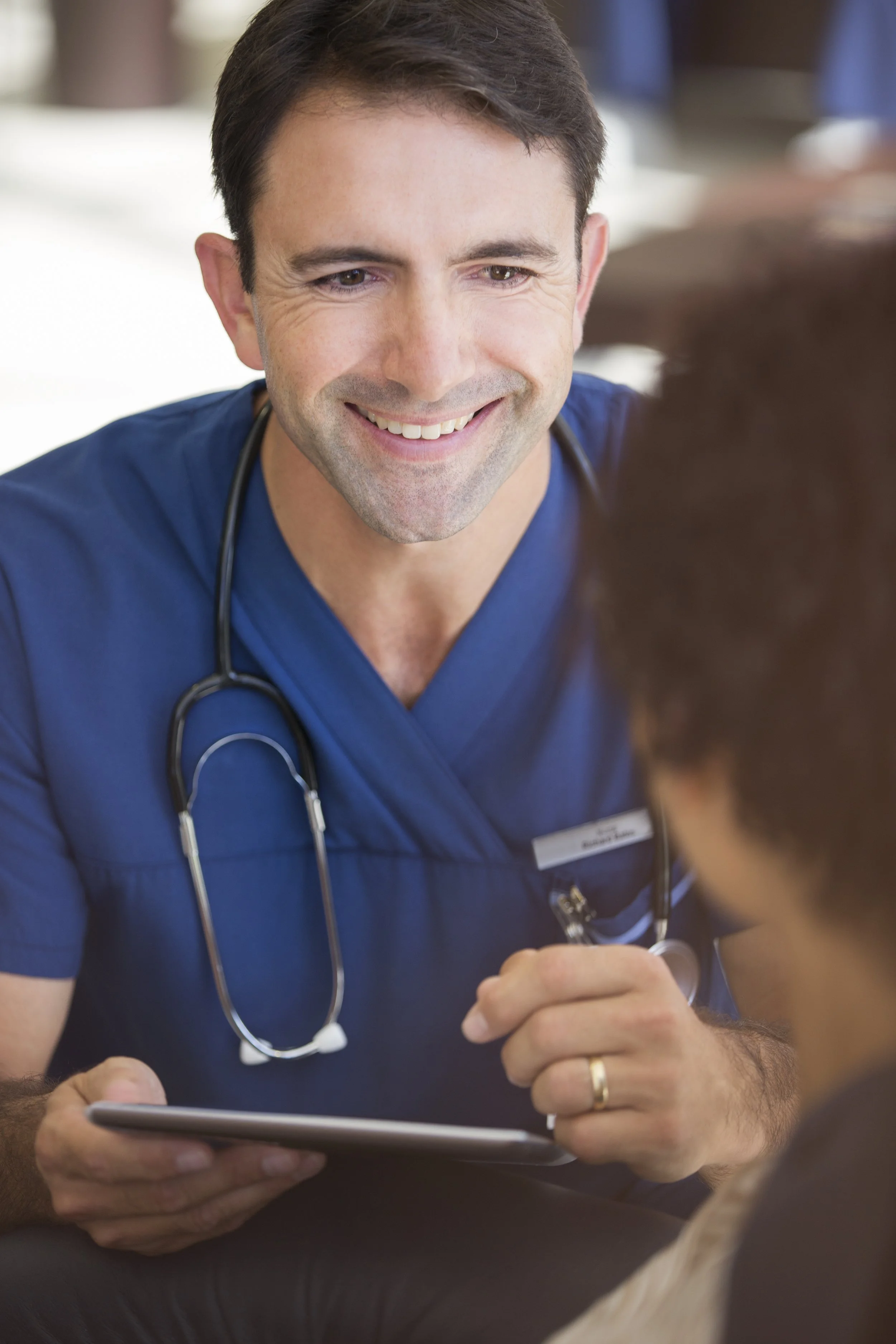 A male healthcare professional with dark hair, wearing blue scrubs and a stethoscope, smiling as he looks at a smartphone, possibly interacting with a patient.