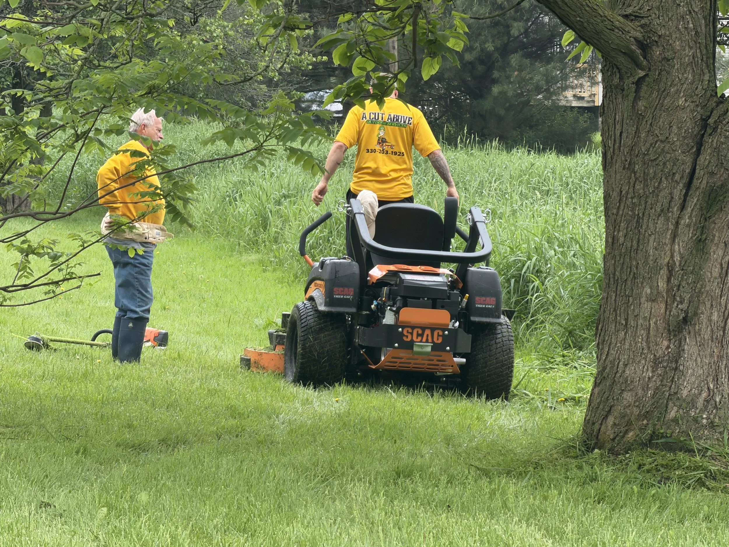 Two men working outdoors in a grassy area surrounded by trees. One man with white hair and dressed in a yellow shirt and blue pants, holding a tool, is talking to another man with tattoos and a yellow shirt that says "A Cut Above Outdoor Services" on the back, pushing a lawn mower.