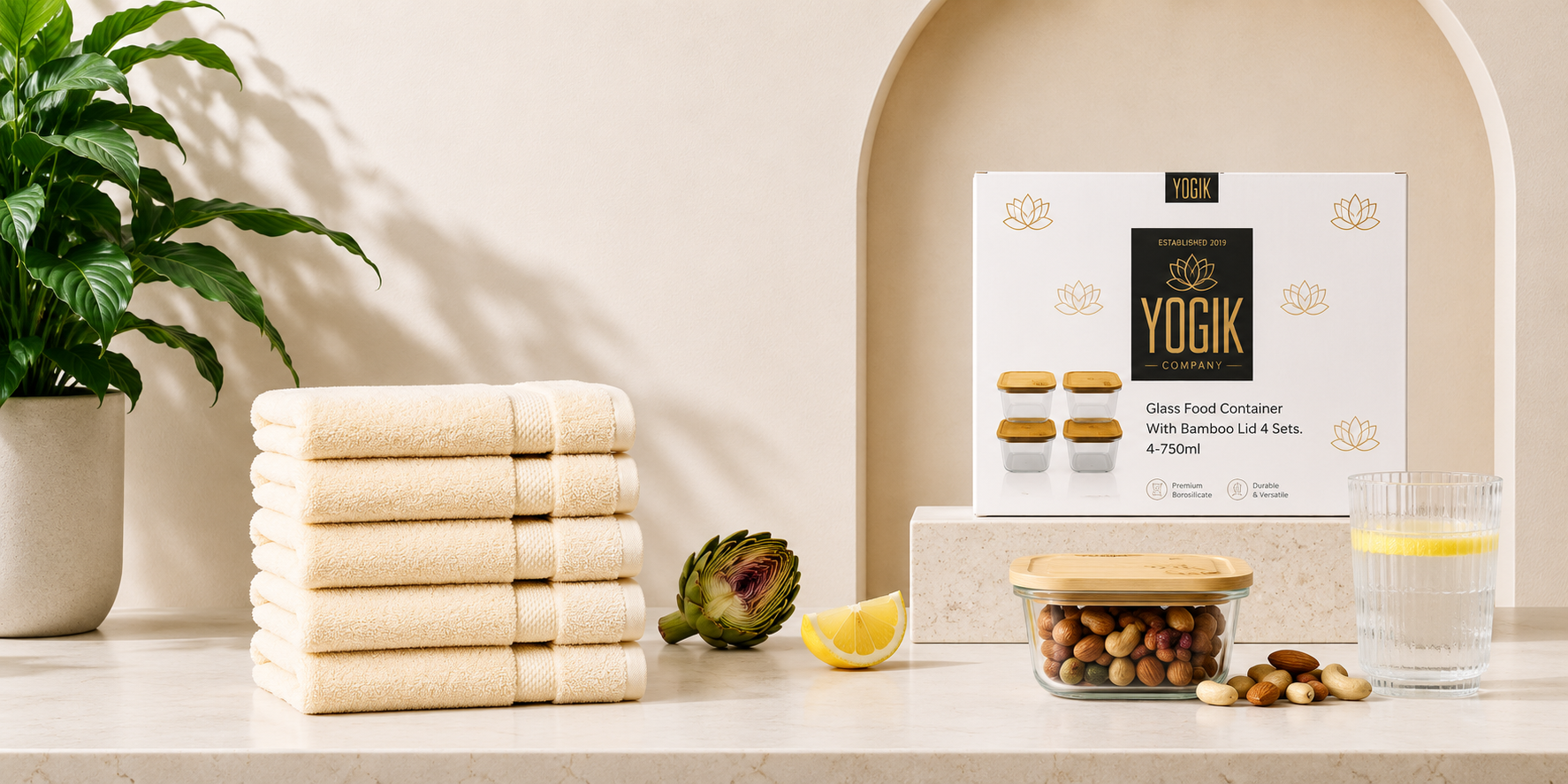 A neatly stacked pile of light beige towels, an artichoke, lemon wedges, a glass of water with lemon, a glass container of mixed nuts with a bamboo lid, and a box of YOGIK glass food containers with bamboo lids on a white surface. There is a potted plant with large green leaves on the left and a decorative arched niche in the background.