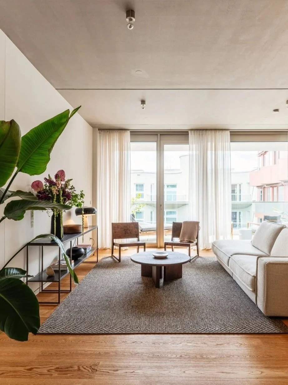 Modernes Wohnzimmer mit großen Fenstern, weißen Vorhängen, einem weißen Sofa, zwei Stühlen, einem runden Couchtisch, einem Teppich, einem Regal mit Dekorationen und einer großen Pflanze.