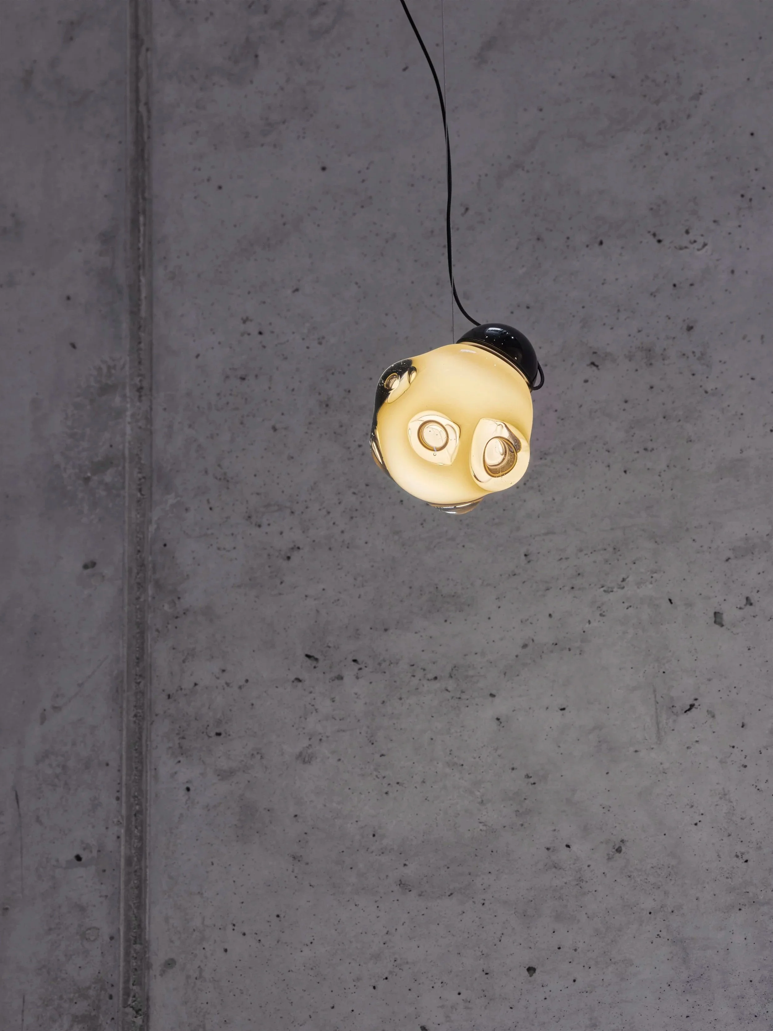 Hängende Lampe mit gelber Glasabdeckung und schwarzen Akzenten, an einer schwarzen Schnur vor einer grauen Betonwand.