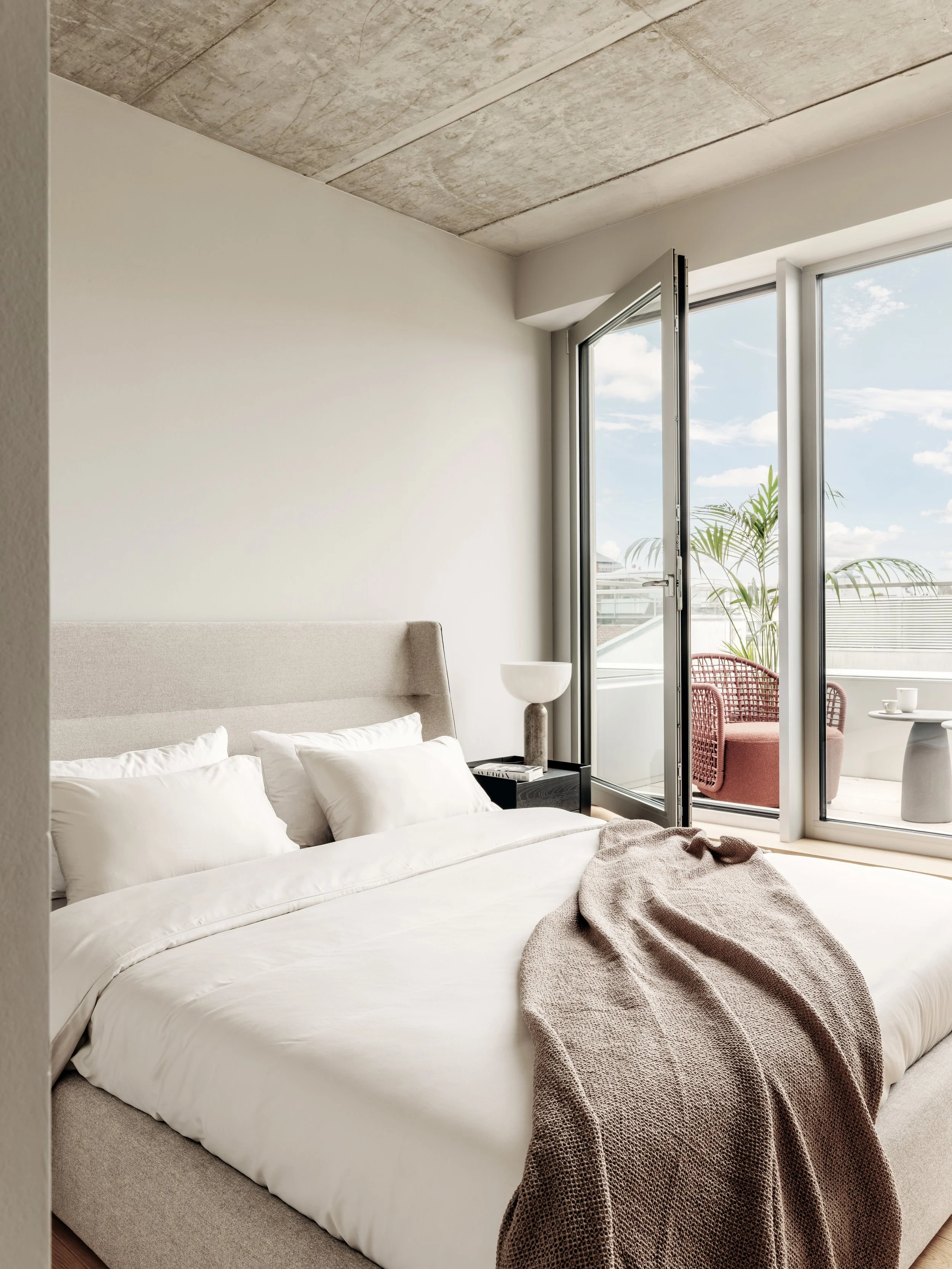 Ein modernes Schlafzimmer mit einer großen, weißen Bettwäsche, mehreren weißen Kissen, einer Nachttischlampe, und einem Balkon mit Sitzgelegenheiten und Pflanzen, durch den man auf den blauen Himmel mit Wolken blickt.