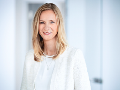 Porträt einer lächelnden Frau mit blondem Haar, in einem weißem Blazer, im Hintergrund ein Büro.