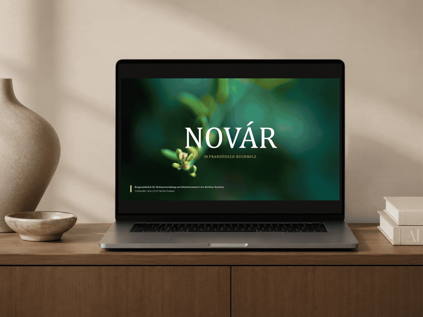 Laptop auf einem Holzschreibtisch mit einem Bildschirmschoner, der das Wort "NOVÁR" in Großbuchstaben zeigt, mit einem grünen, natürlichen Hintergrund. Links befindet sich eine große, beige Vase, daneben eine kleine Teller. Rechts liegen Bücher.