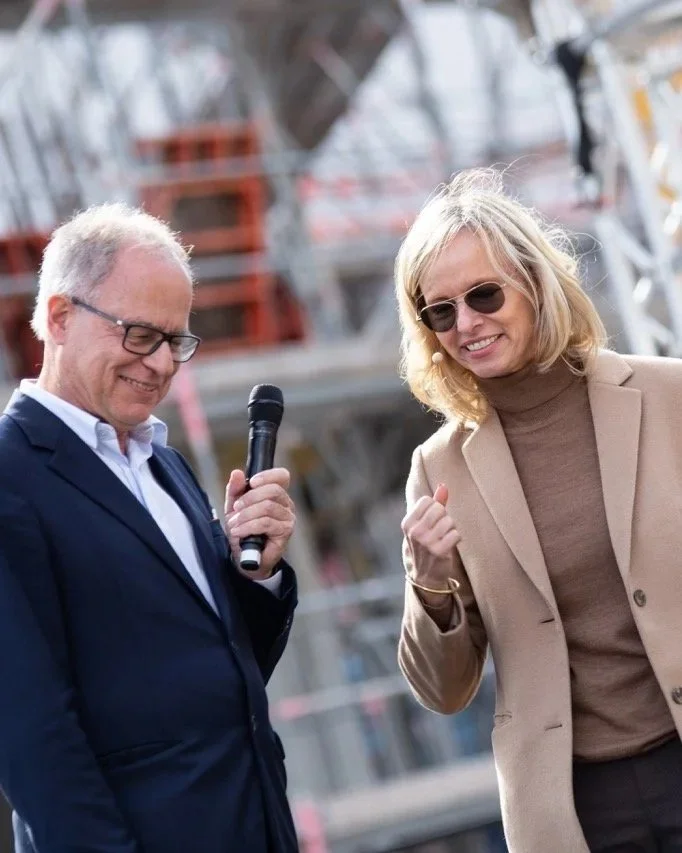 Zwei Menschen, ein Mann im Anzug mit Brille und eine Frau mit Sonnenbrille und beige Jacke, lachen und sprechen bei einer Veranstaltung im Freien.