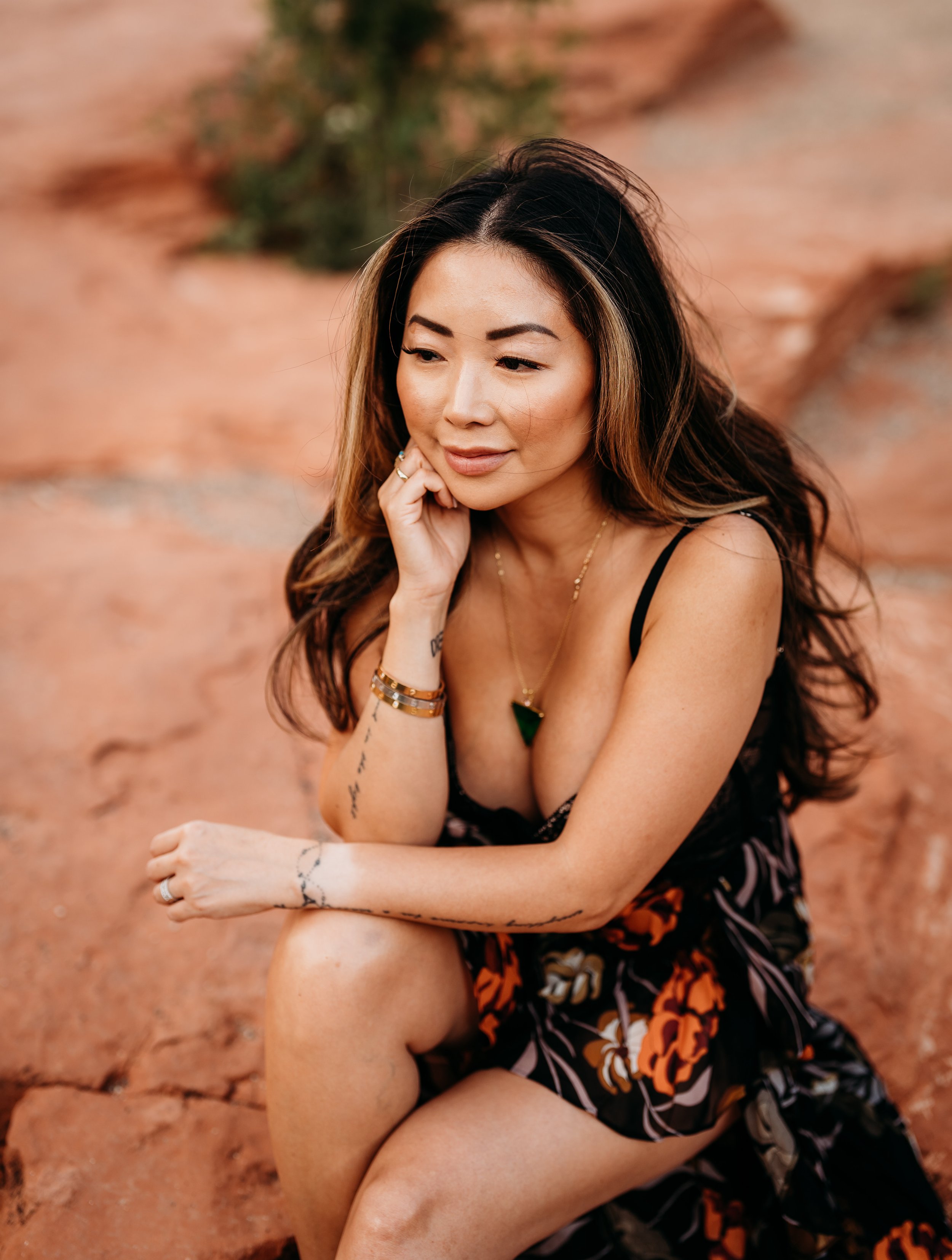 A woman sitting outdoors on red ground, resting her chin on her hand, wearing a black floral dress, with long brown hair and tattoos on her arms.