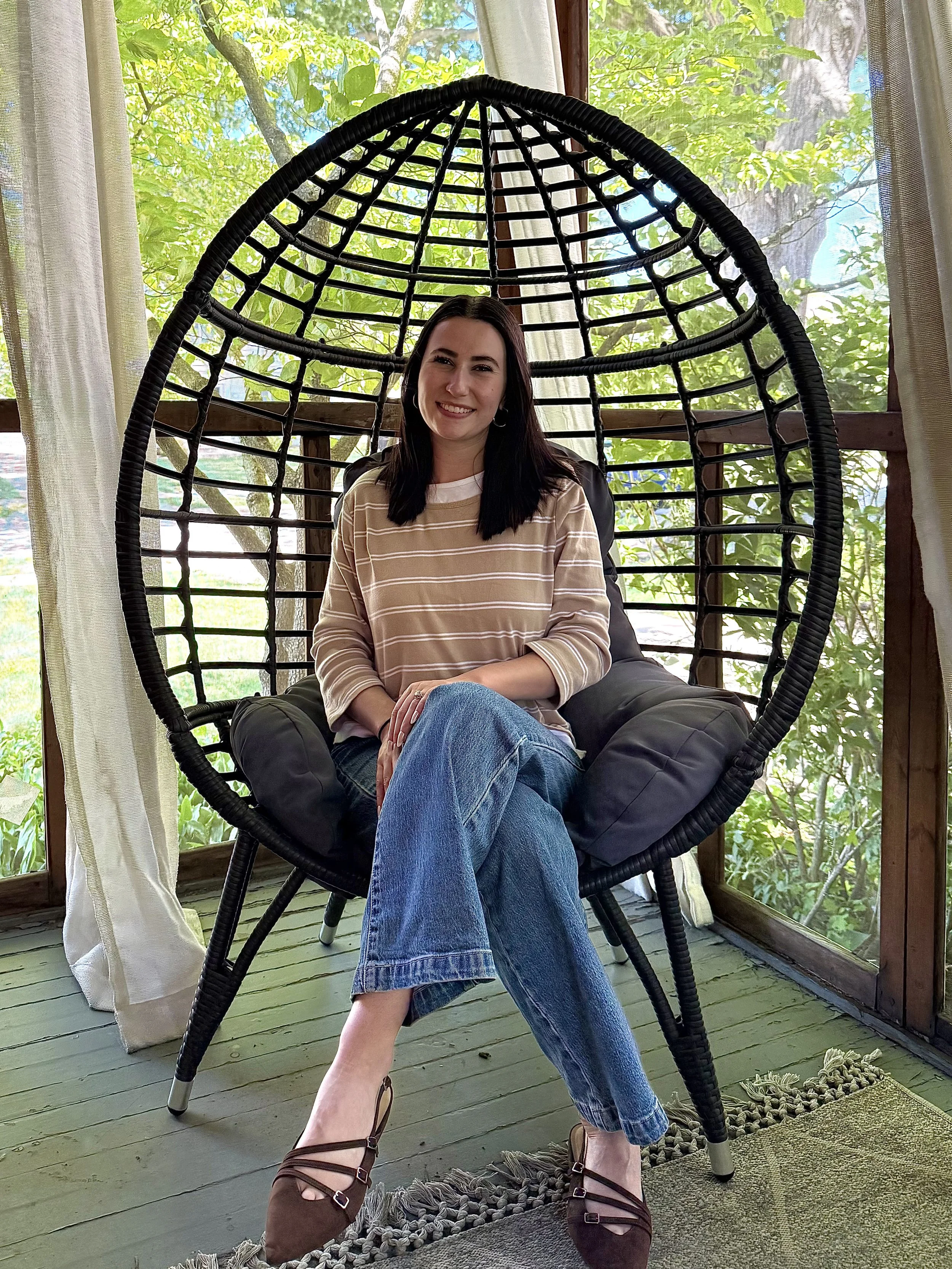 A woman sitting in a hanging chair with a black woven frame, smiling, with a background of lush green trees and sunlight streaming through windows, wearing a beige striped long-sleeve shirt and jeans.