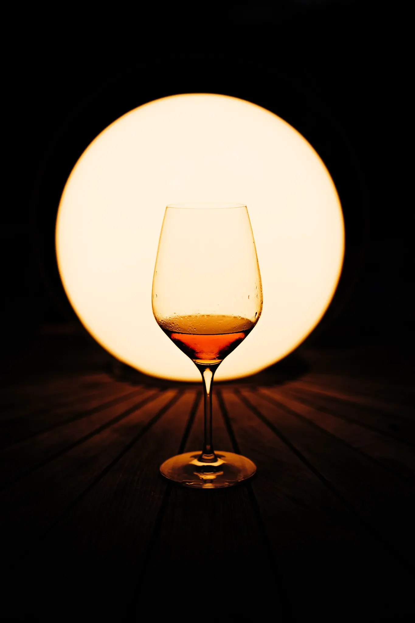 A wine glass filled with red wine in front of a large circular light source on a dark background, casting a shadow on a wooden surface.