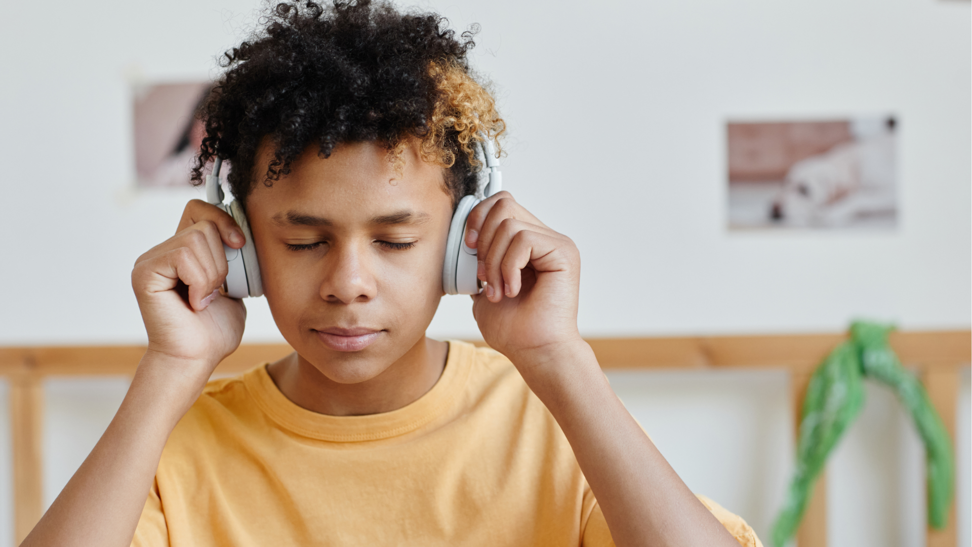 A young person with curly hair wearing a yellow shirt and headphones, with eyes closed, in a room with a white wall and photographs or artwork in the background.