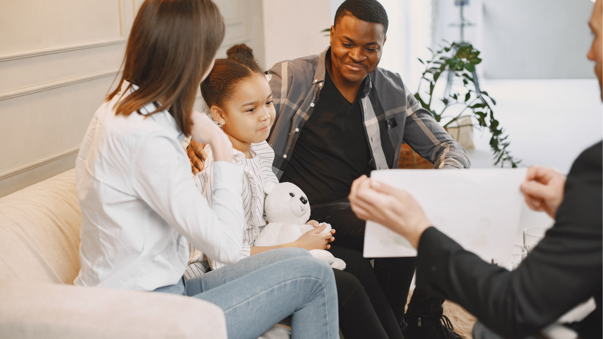 A group of five people, including a young girl with a teddy bear, sitting on a beige couch and participating in a discussion or consultation with a person holding a photo or document in an indoor setting.