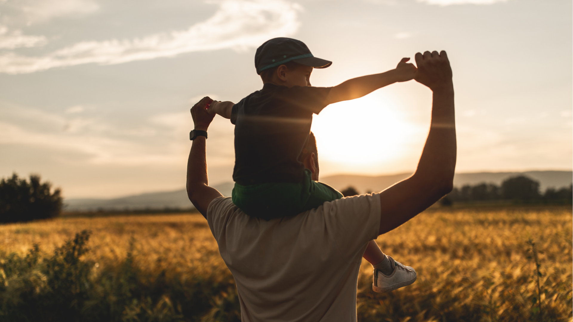 Man lifting child on shoulders at sunset in a field with a scenic distant landscape.