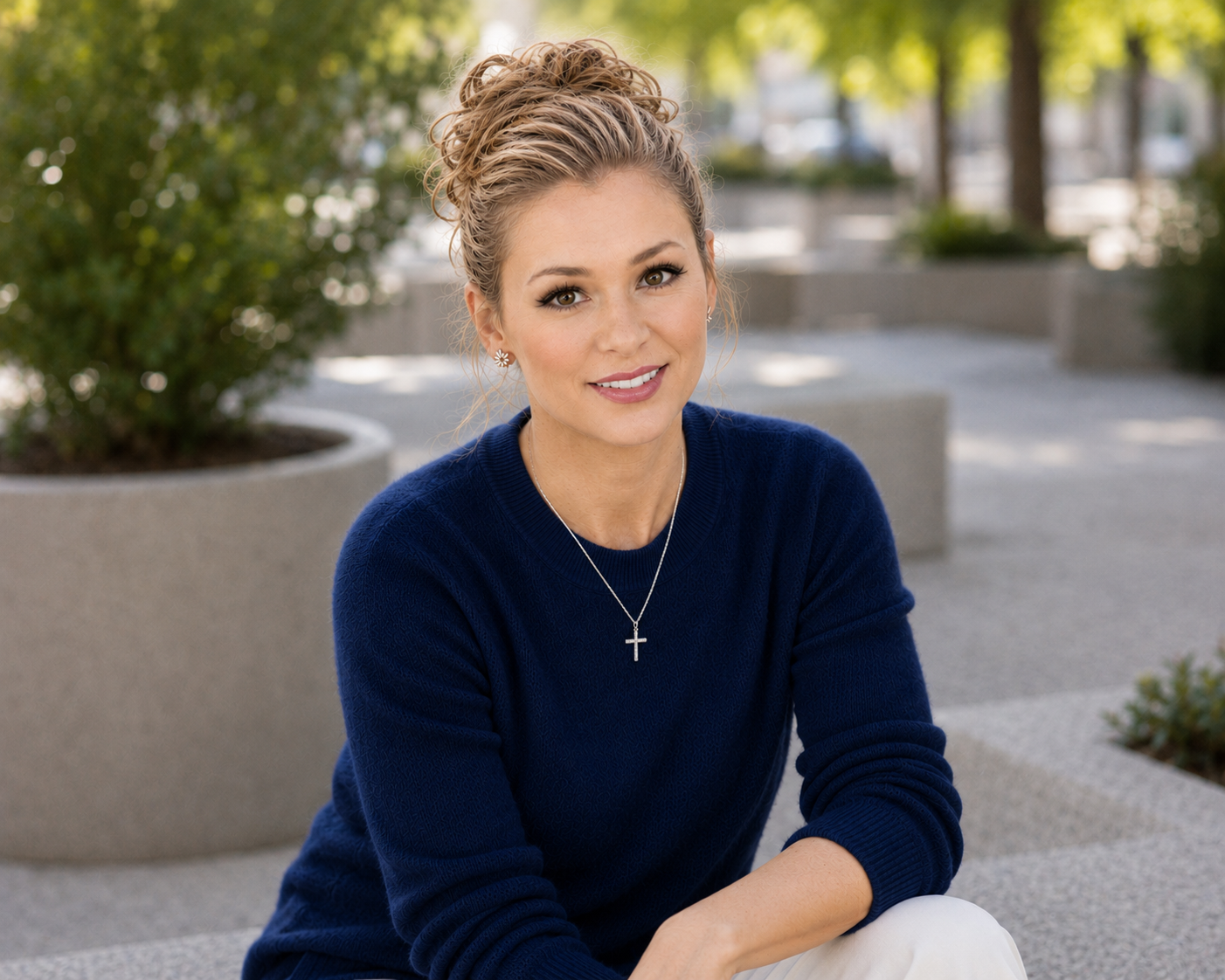 A woman with curly blonde hair, wearing a navy blue sweater and cross necklace, sitting outdoors on a concrete bench with large potted plants and trees in the background.