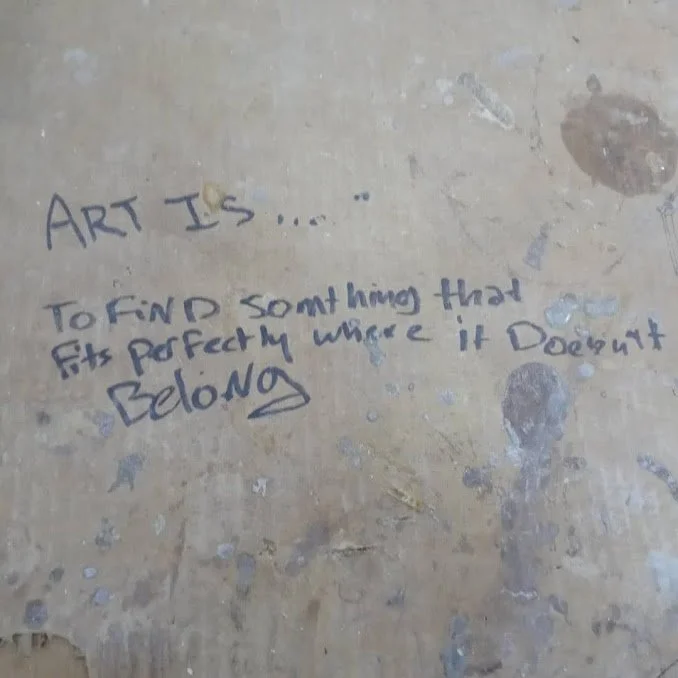Handwritten quote on a worn concrete surface that says, 'ART IS ... TO FIND something that fits perfectly where it doesn't belong'.
