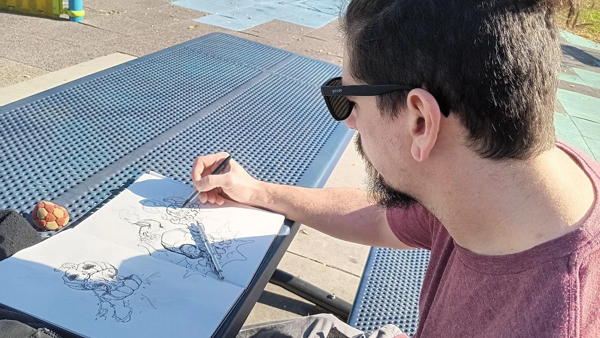 Alex, sitting at outdoor table, drawing comic characters on a large white paper with markers