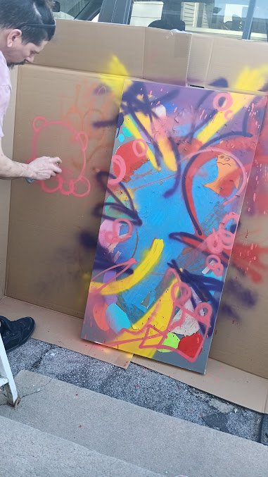 Alex spray painting abstract art with bright colors including pink, yellow, blue, purple, and red on a large canvas in an outdoor setting.