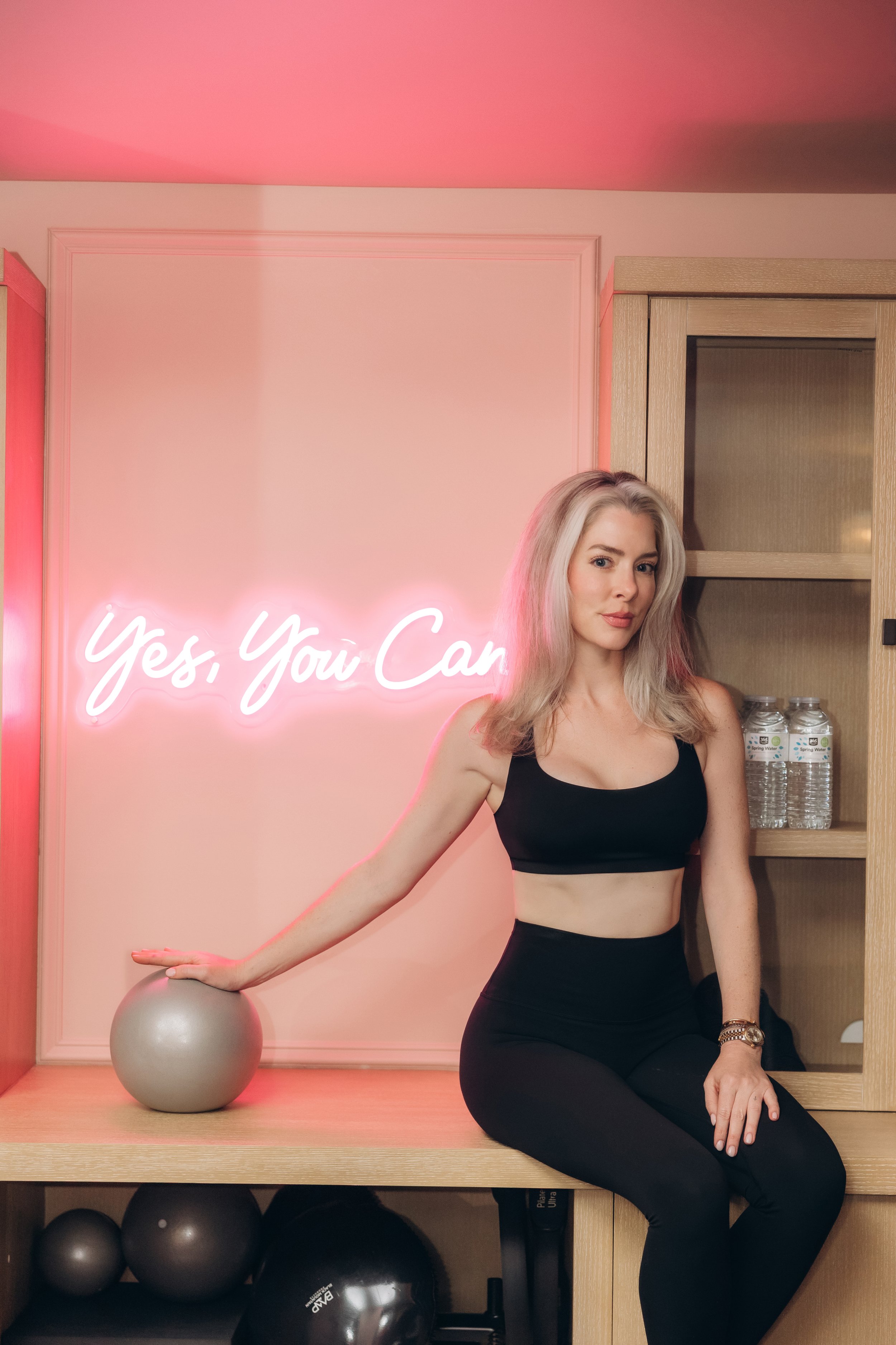 A woman with platinum blonde hair, wearing a black sports bra and leggings, sitting on a wooden bench with a pink wall behind her. To her left, a neon sign reads 'Yes, You Can'. There are water bottles on a shelf behind her, and exercise equipment, including yoga balls, on the shelf below.