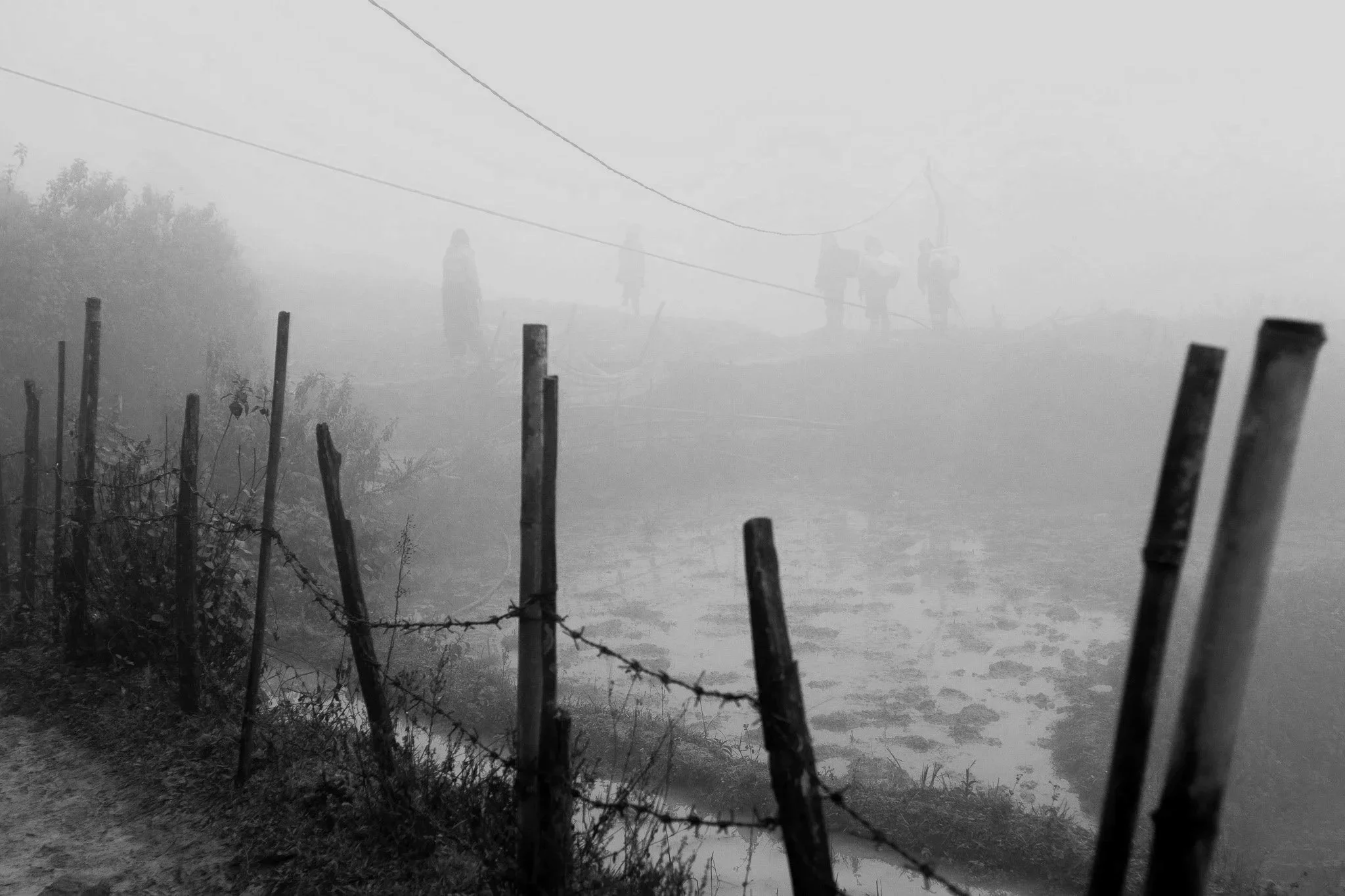 A black-and-white photo of a foggy outdoor scene with silhouettes of four people walking in the distance, seen through a barbed-wire fence near a body of water.