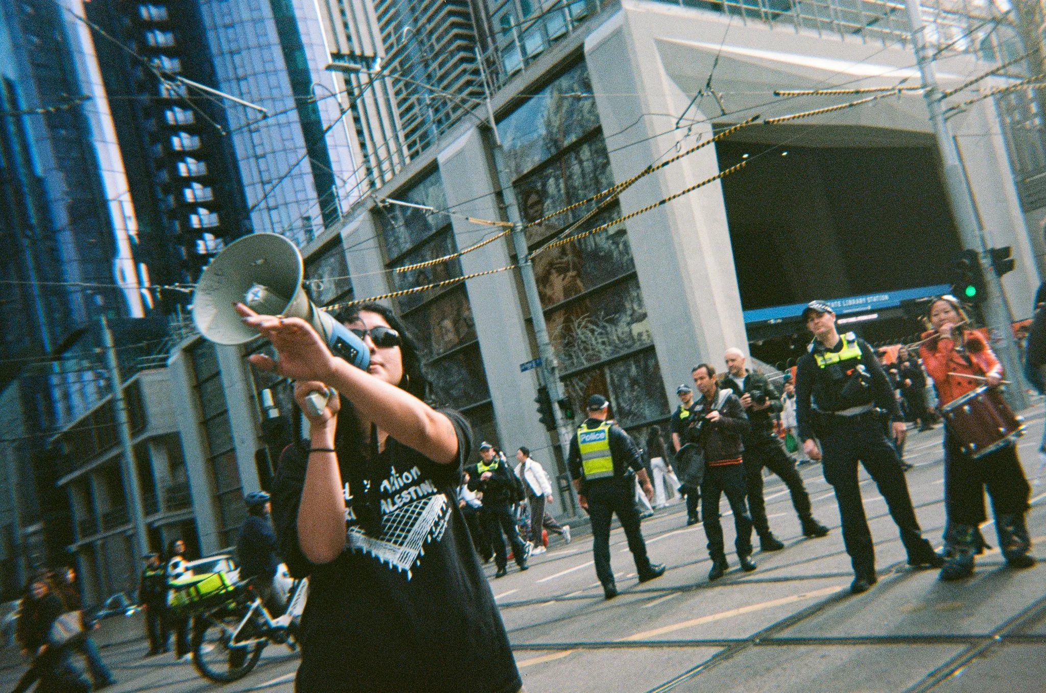 A woman with sunglasses holding a megaphone at a busy urban protest with police officers and pedestrians in the background.