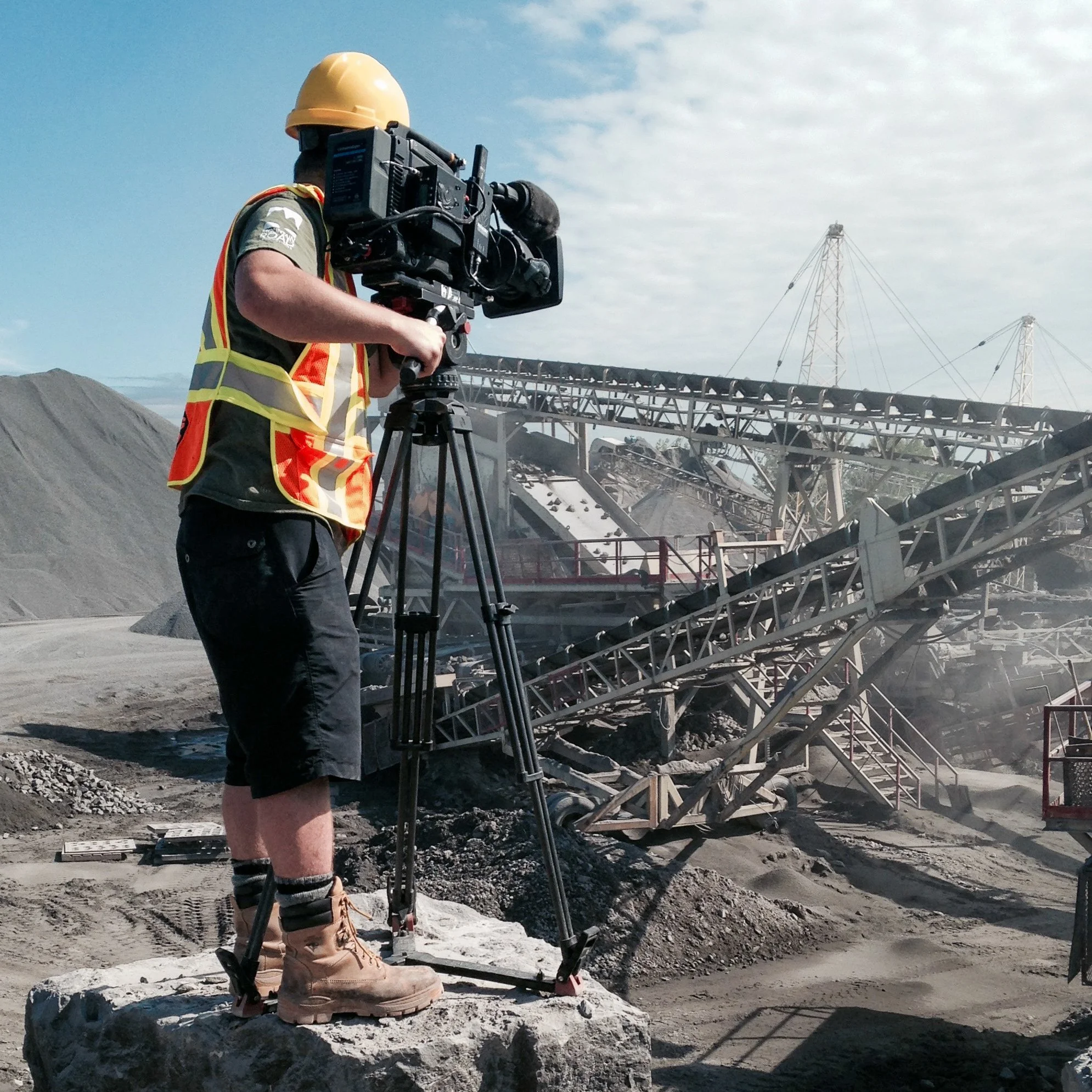 Person in safety gear operating a camera on a tripod at an industrial site with conveyor belts and power lines in the background.
