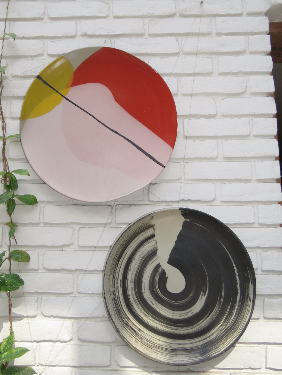 Two abstract painted ceramic plates mounted on a white brick wall, with one plate above the other. The top plate has sections of red, yellow, pink, and black, while the bottom plate has black and white concentric circular patterns.