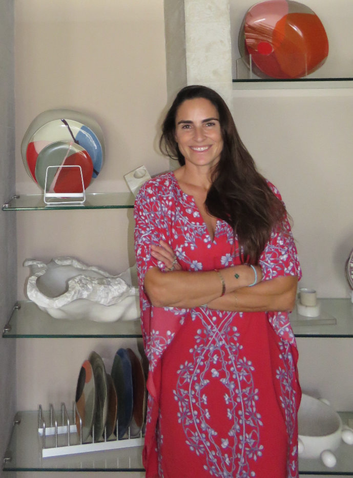 A woman with long dark hair wearing a red dress with light blue floral embroidery, standing with her arms crossed and smiling in front of glass shelves with decorative art pieces, including colorful glass bowls and abstract sculptures.