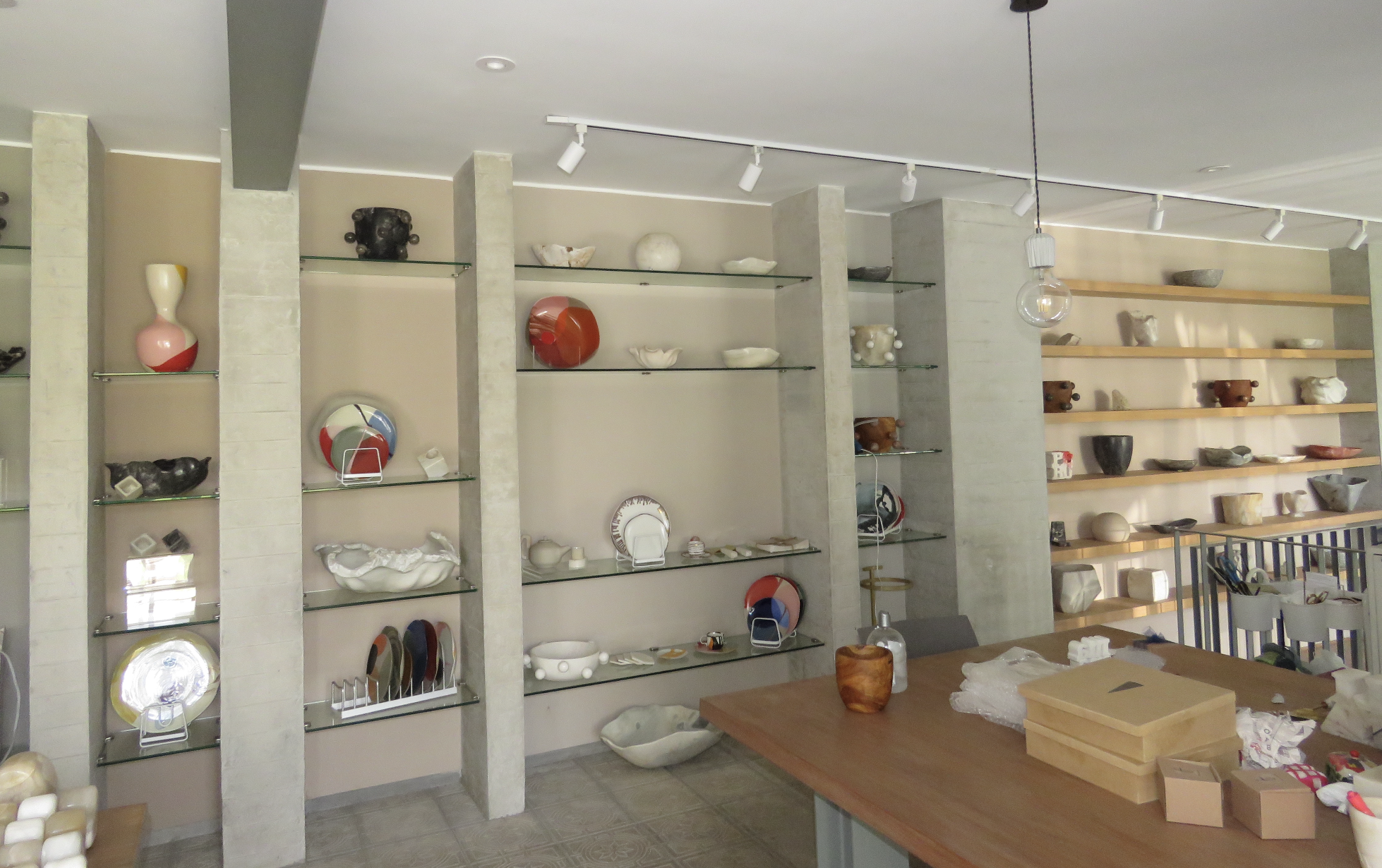 Interior of a store with shelves displaying ceramic vases, bowls, and decorative items. A wooden table with a box and other packaging materials is in the foreground. Track lighting fixtures are mounted on the ceiling.