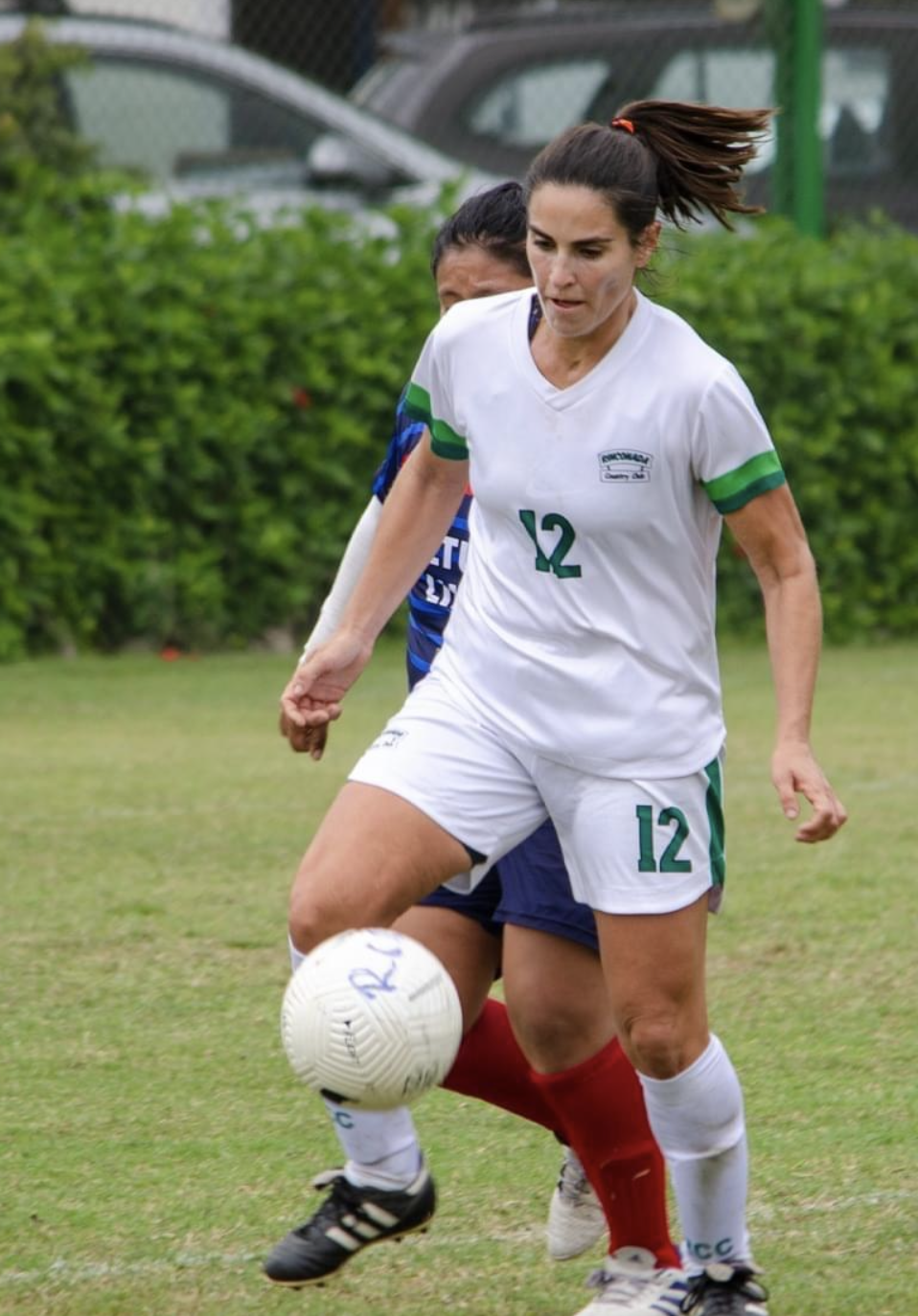 Female soccer players competing for the ball on the field, with one in a white uniform numbered 12 and the other in a blue and red uniform.