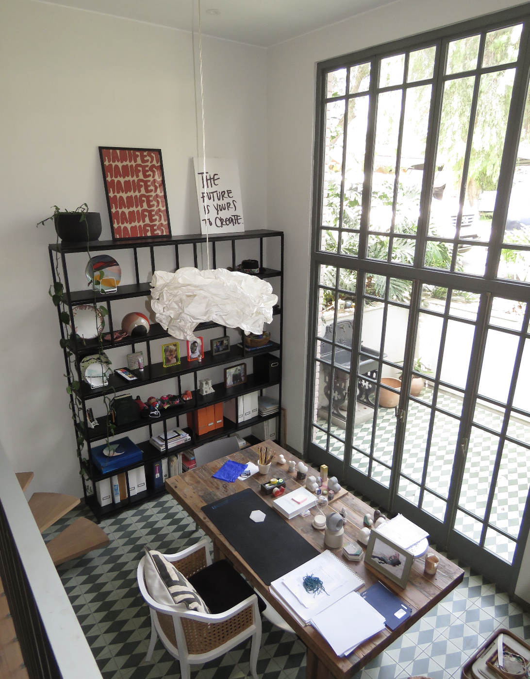A home office with a wooden desk, white chair, black shelving unit, and large window with black grid frames.