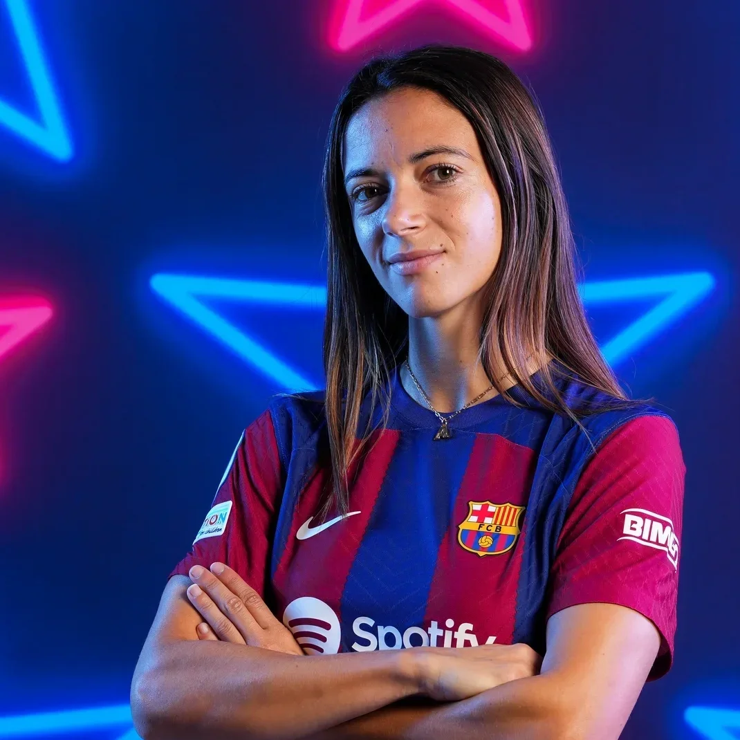 A woman wearing a Barcelona football jersey standing against a dark background with pink and blue neon star lights. She has long brown hair and is crossing her arms.